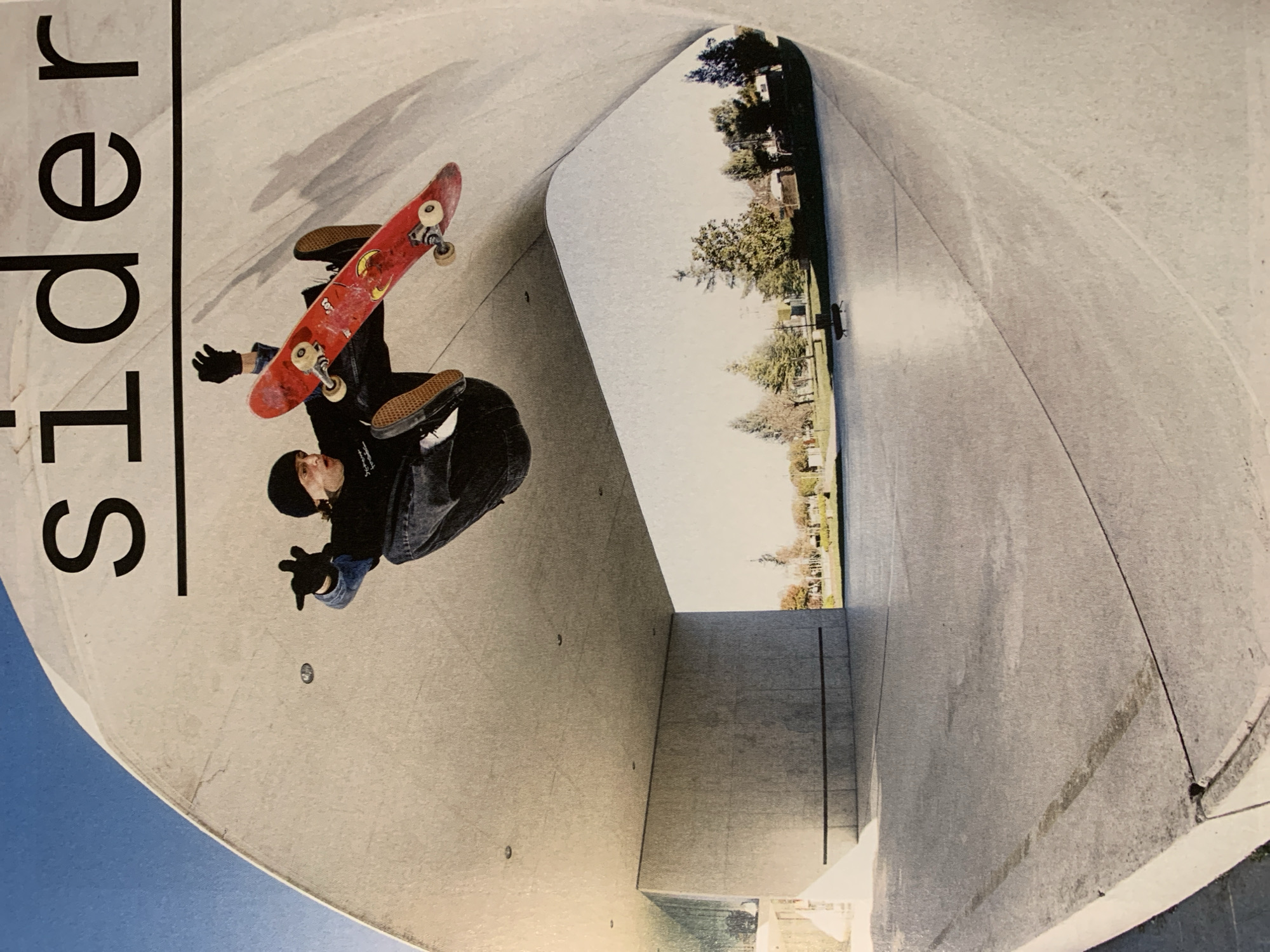 A skateboarder is performing a trick inside a concrete skatepark structure, likely a quarter pipe or a full pipe, with a view of trees and buildings visible through the opening. The skateboarder is wearing a black beanie, a dark jacket, jeans, and Vans shoes and has black gloves on. They are oriented upside down in the frame, with the red skateboard above them. The word "Slider" is partially visible on the left side. Given the style of architecture and layout, the location is likely the Skatepark Mar Bella in Barcelona, Spain, but the exact location and certainty cannot be determined solely from this image. The photo appears to be a snapshot from a magazine or similar publication.