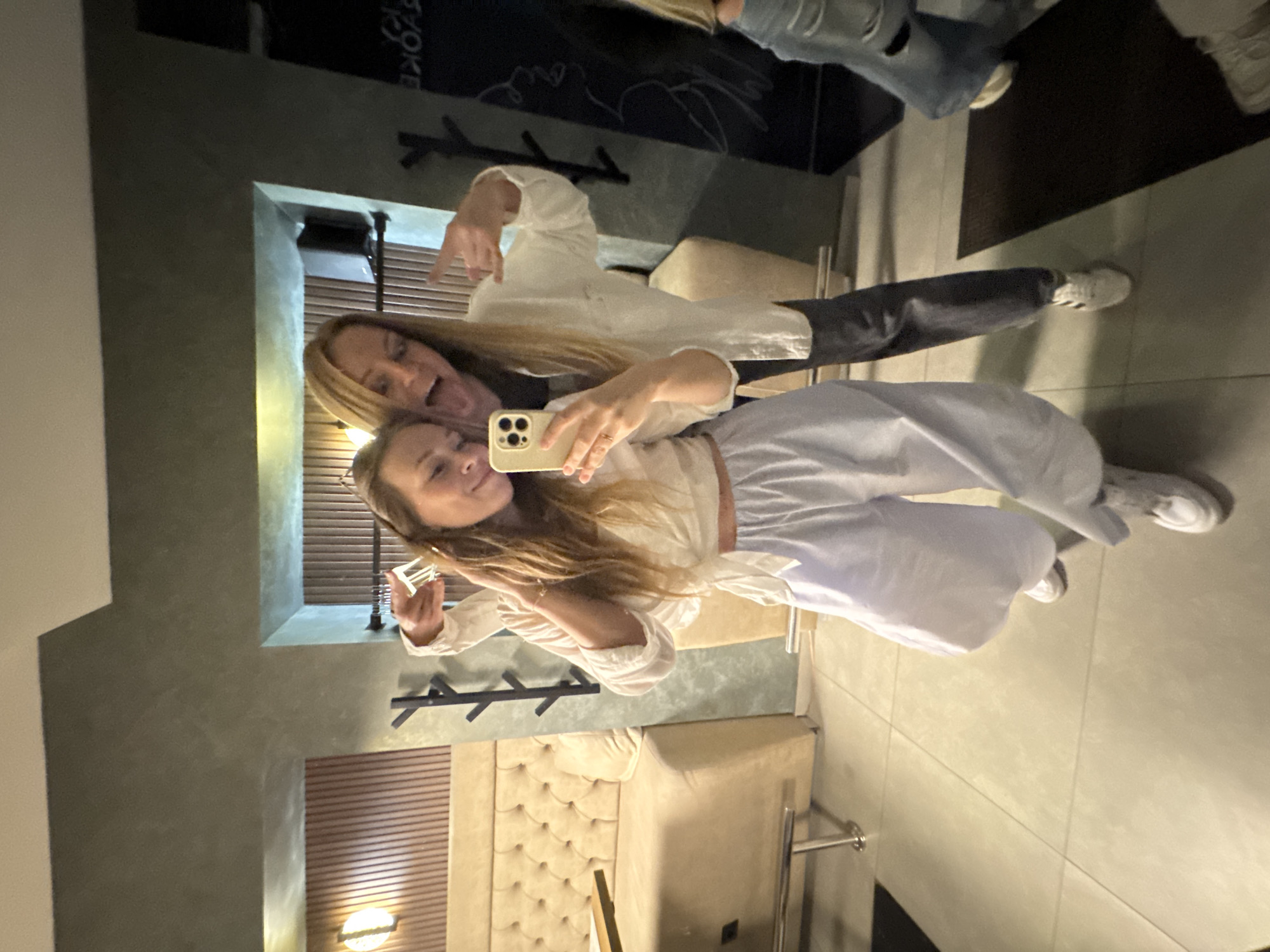 Two women are taking a selfie indoors. One woman has long blonde hair and is wearing a white top, dark jeans, and white sneakers. She is making a hand gesture. The other woman is in the foreground, also with long blonde hair, wearing a white top, loose white pants, and white sneakers. She is holding a cell phone up to take the picture. Behind them is a beige-colored wall and a light fixture. The location may be in Budva, Montenegro, but that cannot be determined definitively from the image.