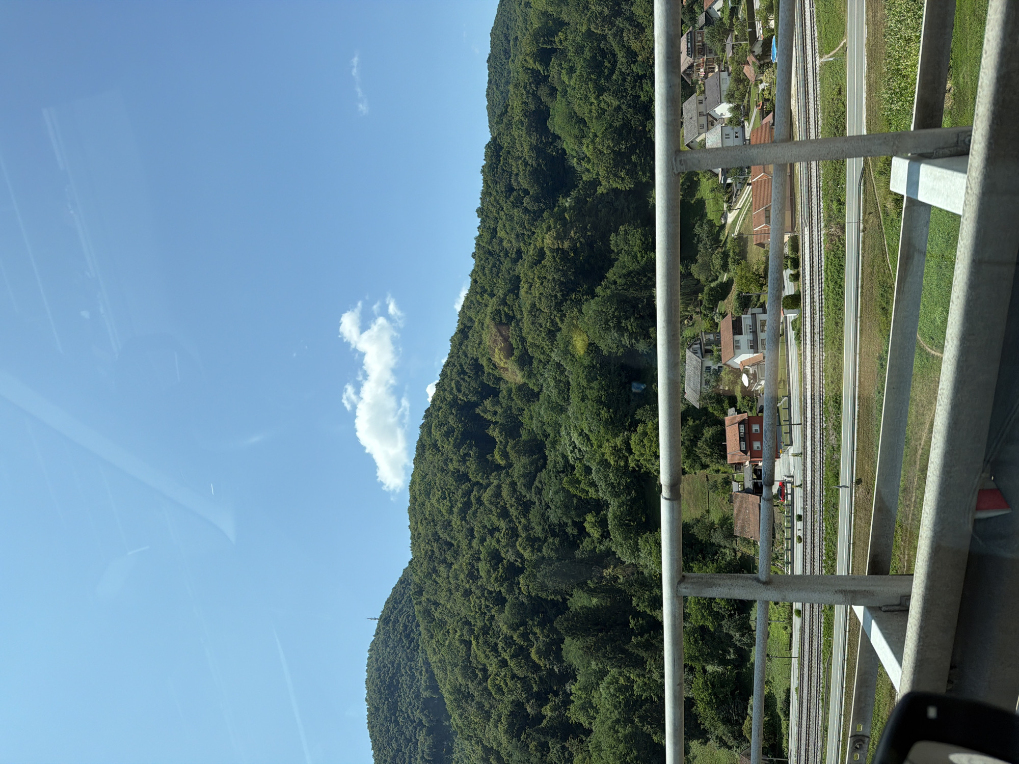 The image captures a view from a railing overlooking a rural landscape in Ravninsko, Croatia. The foreground features a metal railing and a portion of a car's side mirror. Beyond the railing, a road and train tracks are visible, separating the foreground from a village with several houses. In the background, a lush, tree-covered mountain dominates the scene under a bright blue sky with scattered white clouds.