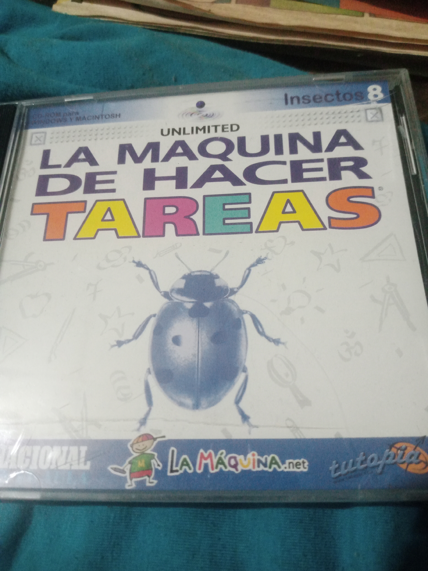 The image shows a CD-ROM game case for "Insectos 8" with the title "LA MAQUINA DE HACER TAREAS" which translates to "The Machine for Doing Homework." The case features a large illustration of a blue beetle on a white background with various educational icons like stars, planets, and rulers subtly imprinted. In the lower portion of the case, the text "NACIONAL" is visible, along with a cartoon drawing of a child and the website "LA MAQUINA.net tutapia." The CD case is positioned on a teal-colored fabric surface, which appears to be part of a bedspread or blanket. The background suggests other stacked items, possibly books or more media. The lighting is dim and appears to be coming from above and to the right. There is no indication or information within the image that places this scene in Santa Ana, Venezuela, or any specific city.