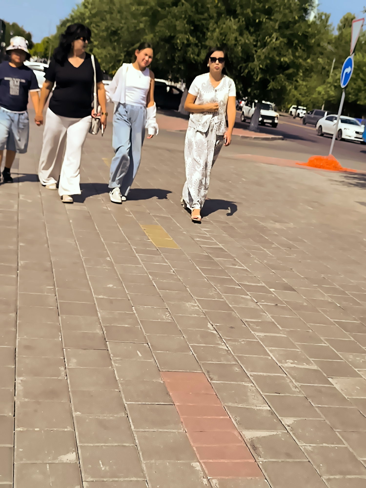 Four people are walking on a sidewalk in front of a street with some cars. The sidewalk is paved with brown rectangular tiles. On the left, a child wearing a dark blue t-shirt, light blue shorts, and a white bucket hat is walking. Next to the child, a woman with dark hair, sunglasses, and a black v-neck shirt with white pants is walking and holding hands with the child. To their right, a young woman in a white tank top, white unbuttoned shirt, and light blue jeans is walking. The furthest person on the right is a woman wearing a white printed outfit and sunglasses, walking with her hands in her pockets. In the background, trees line the street, and several cars, including a white SUV and a white sedan, are visible on the road. A blue and white directional sign is also present. The scene appears to be in Qarağandı city administration, Kazakhstan, under a bright, sunny sky.