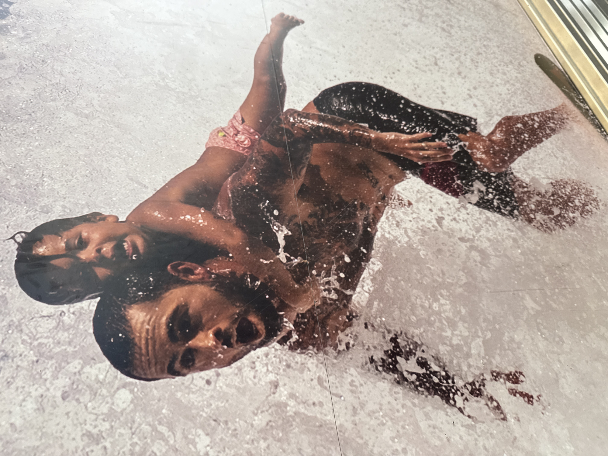 An energetic, action-shot photograph captures two individuals, an adult and a child, actively playing and splashing in water. The adult, appearing to be a man with dark hair and stubble, is positioned largely submerged, facing upwards with an open-mouthed smile, suggesting excitement or a laugh. The child, with dark hair and wearing pink patterned swim bottoms, is on the adult's back, also facing upwards with a broad, joyful smile.
Both subjects are covered in water droplets and surrounded by turbulent white splashes and foam, indicating a dynamic moment in an aquatic setting, possibly a water slide or vigorous play. The adult wears dark swim trunks. The background consists almost entirely of rushing, white water, with no specific landmarks visible to determine the location beyond it being an outdoor water activity. The lighting is bright and even, consistent with daytime. The overall emotion conveyed is one of joy, fun, and exhilaration. There is no visible text in the image.