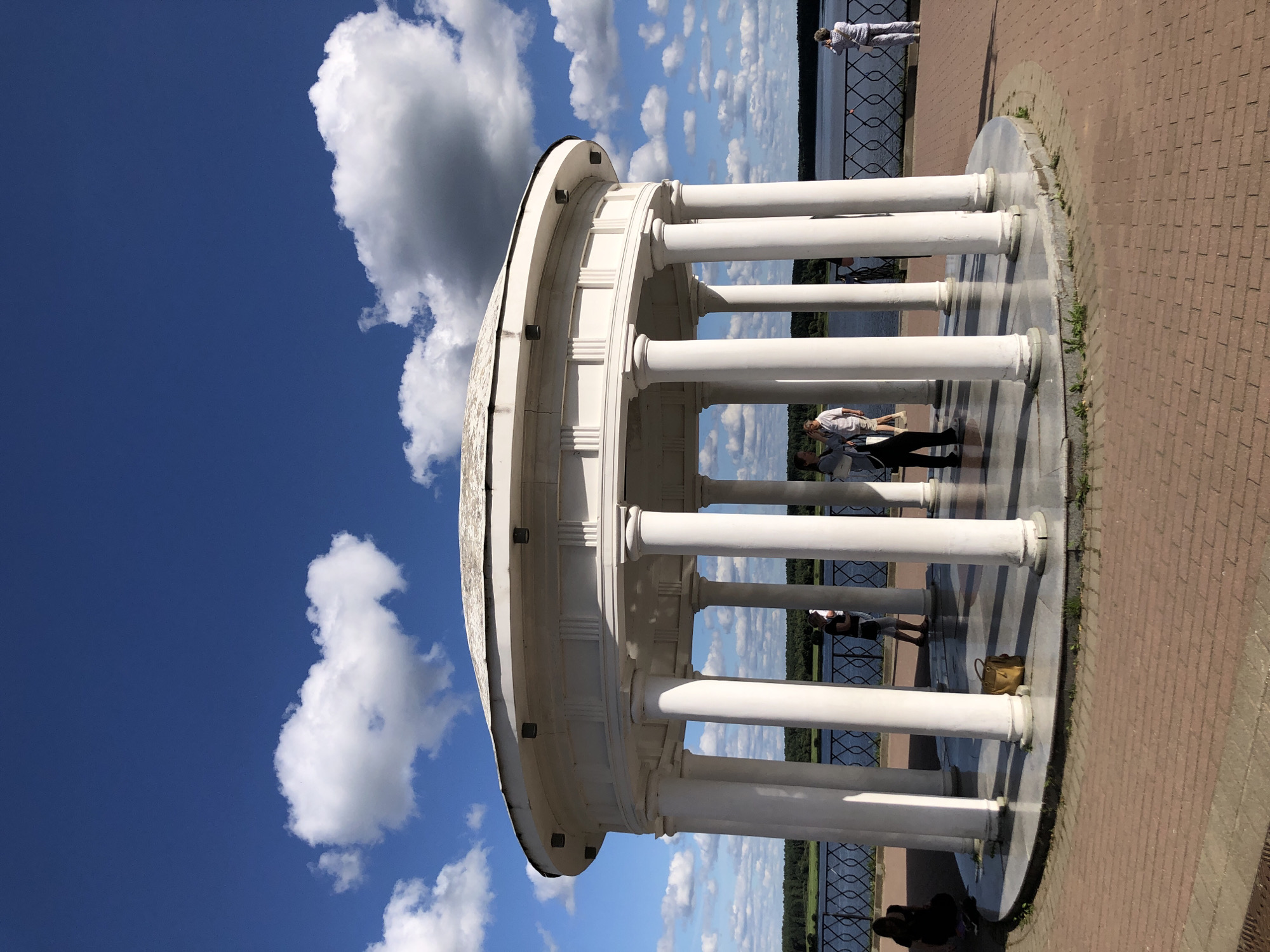 The image shows a white, classical-style rotunda with a domed roof and multiple columns, situated on a paved circular platform. A body of water, likely a river, is visible in the background, bordered by a dark railing and a distant tree line. The sky is a bright blue with scattered white and gray clouds. Several people are present: two individuals are standing within the rotunda, one appearing to converse and the other looking down; a woman is crouched on the ground near the rotunda's edge; another person is visible in the distance, standing by the railing and facing away from the camera; and another individual is standing near the rotunda's entrance. A yellow bag rests on the platform inside the rotunda. The overall scene suggests a public park or waterfront area. The location is identified as the Rotunda of Love in Myshkin, Russia.
