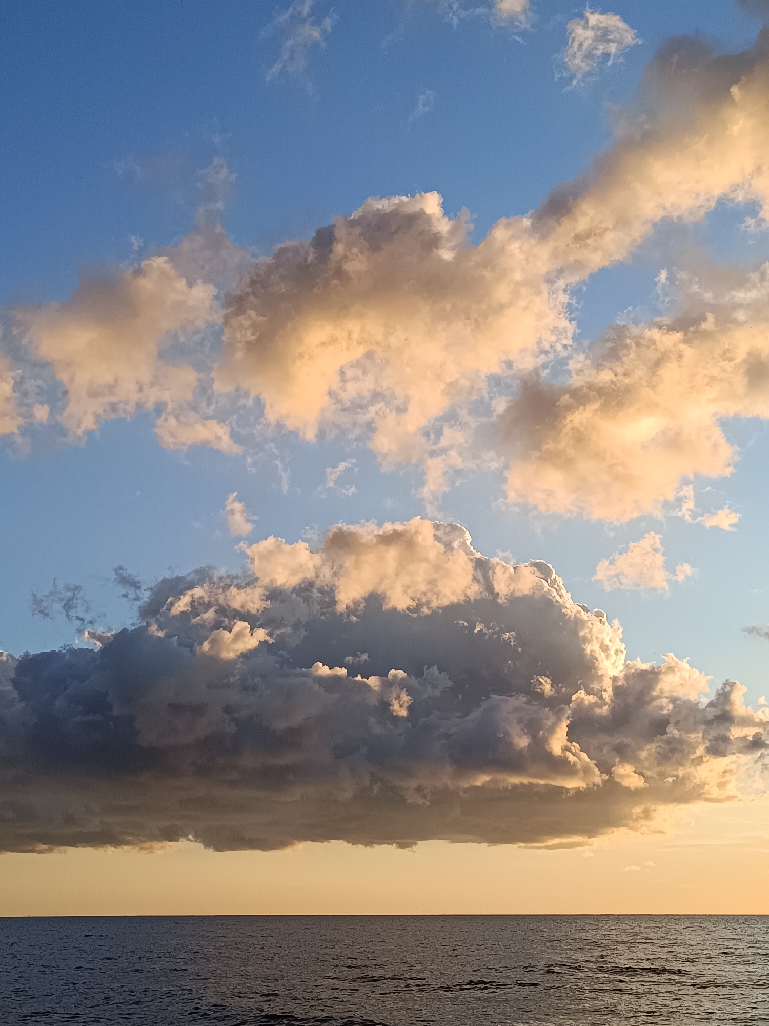 A wide shot captures a tranquil scene of the sea meeting the sky. The sea is a deep, dark blue, with subtle ripples on its surface. Above, the sky transitions from a pale yellow hue near the horizon to a vibrant blue. Large, dramatic clouds dominate the upper portion of the image. Some clouds are illuminated with warm, golden light, suggesting either sunrise or sunset, while others are shaded in darker tones of grey and white. The composition is divided horizontally, with the sea occupying the bottom third and the sky and clouds filling the remaining two-thirds. There are no people or man-made objects visible in the frame, creating a sense of natural serenity and vastness. The location is described as Unknown, Unknown.