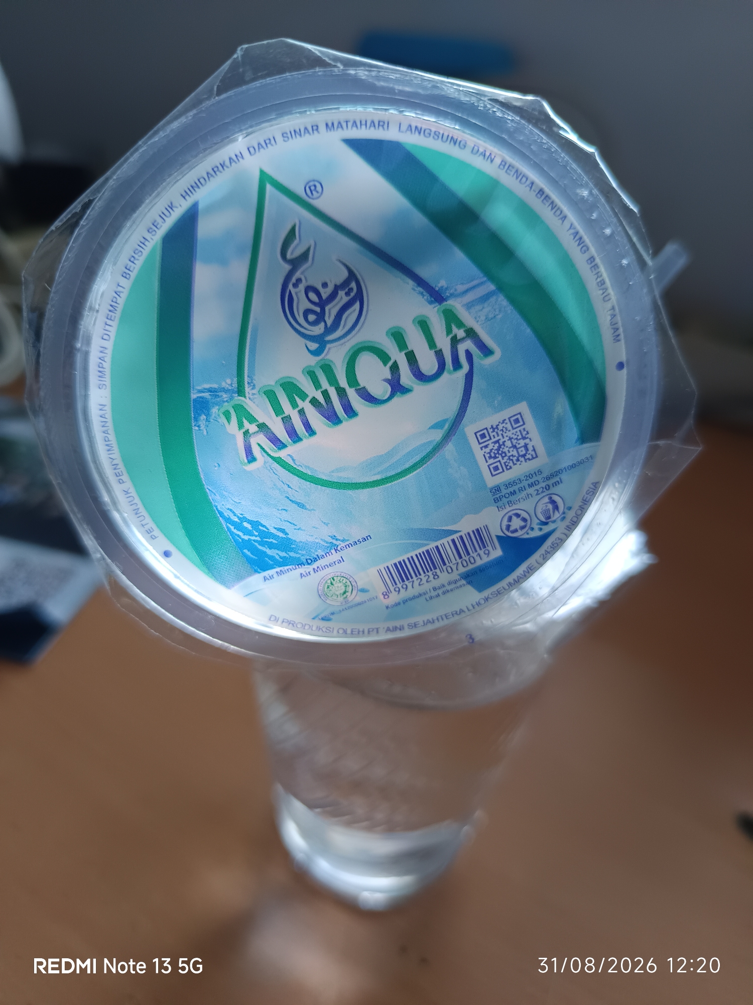 The image shows a sealed plastic cup of mineral water, likely Aqua brand, placed on a wooden surface. The lid of the cup has a blue and green design with the brand name "AQUA" prominently displayed in the center. Text on the lid indicates storage instructions in Indonesian, such as "SIMPAN DITEMPAT BERSIH, SEJUK, HINDARKAN DARI SINAR MATAHARI LANGSUNG DAN BENDA-BENDA YANG BERBAU TAJAM" (Store in a clean, cool place, avoid direct sunlight and strong-smelling objects). It also includes certifications and product details like "SNI 3553-2015", "BPOM RI MD 265201003031", and "Isi Bersih 220 ml". The cup is partially filled with water. In the bottom left corner, the text "REDMI Note 13 5G" suggests the device used to capture the image. The bottom right corner displays a date and time: "31/08/2026 12:20". The overall environment appears to be indoors, possibly a desk or table. There is no indication of the specific location within the City of Medan, Indonesia.