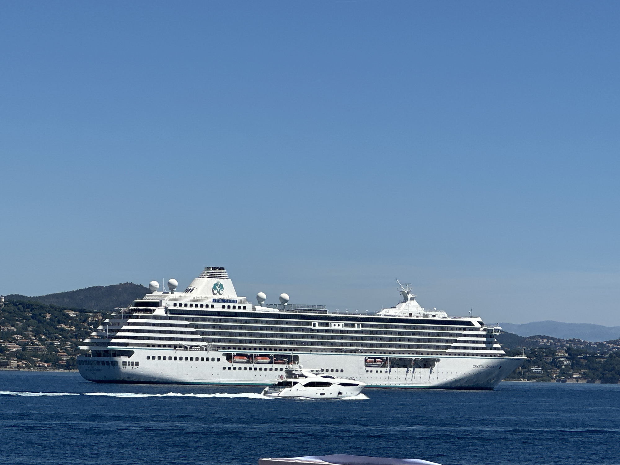 A large white cruise ship, the "Crystal Serenity," is depicted sailing through the blue waters of the Mediterranean Sea off the coast of Saint-Tropez, France. The ship is the dominant subject in the image, taking up a significant portion of the frame. It is oriented to the right, with its bow angled slightly towards the viewer. The ship features multiple decks with numerous windows, and lifeboats are visible along its sides.
In the foreground, a smaller white yacht is also underway, moving from left to right and leaving a white wake in the water. This yacht is positioned in front of the cruise ship.
The background consists of a lush, green, hilly coastline dotted with houses, suggesting a Mediterranean landscape. The sky is clear and bright blue. The scene appears to be captured on a sunny day, with clear visibility. The overall impression is one of a serene maritime setting in a popular coastal resort town.