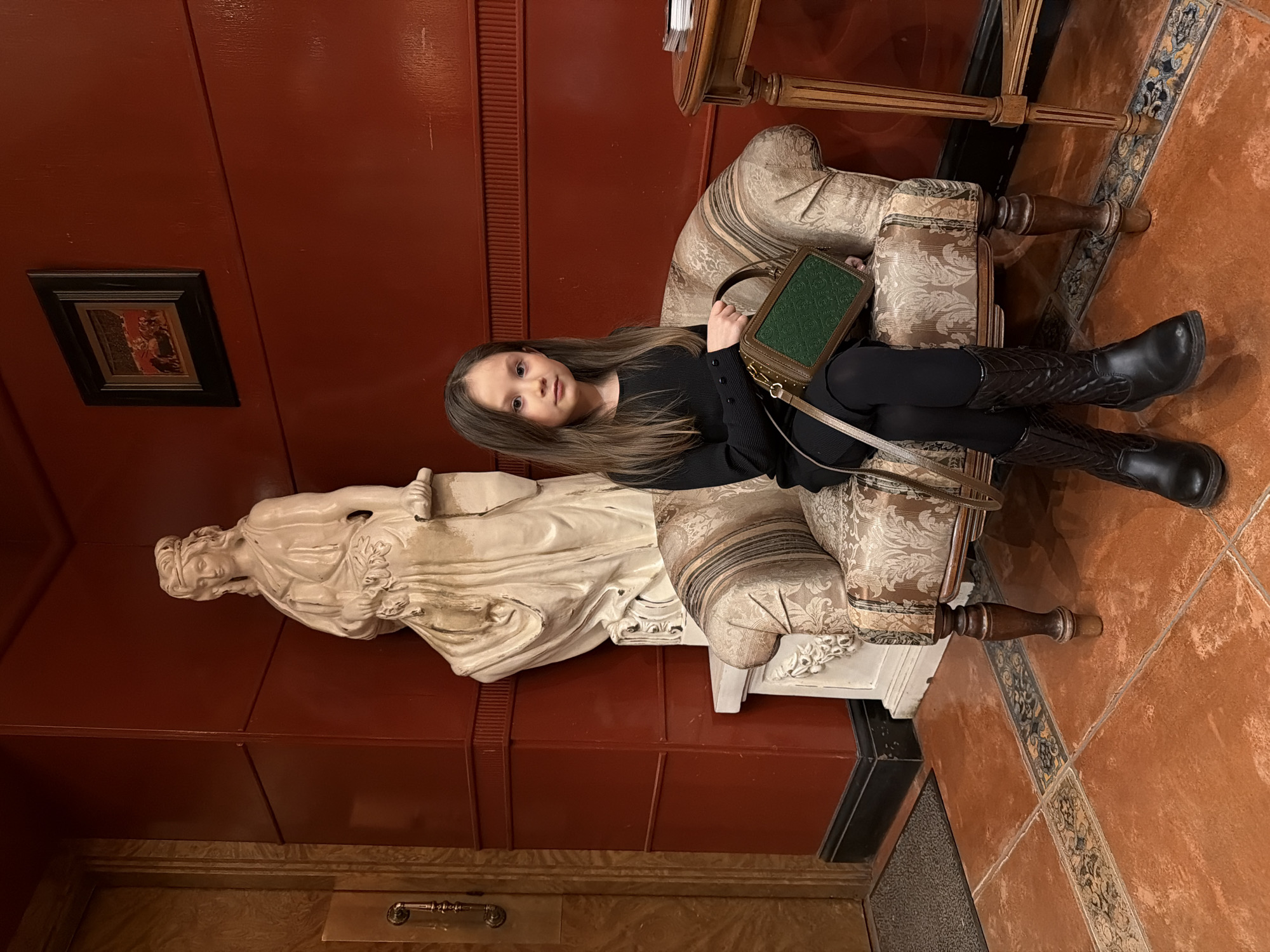 A young girl is seated on an ornate armchair in an interior setting. She is dressed in a black outfit, including leggings and a long-sleeved top, and wears black knee-high boots. She holds a green, rectangular handbag with gold detailing on her lap.
To the left of the girl, a white statue of a draped female figure rests on a plinth against a deep red wall. A framed picture hangs on the wall above the statue.
The armchair is upholstered in a patterned fabric with beige and brown tones. An elaborate side table stands to the upper right of the chair. The floor is tiled in a warm orange hue with decorative borders.
The setting is in Perm, Russia.