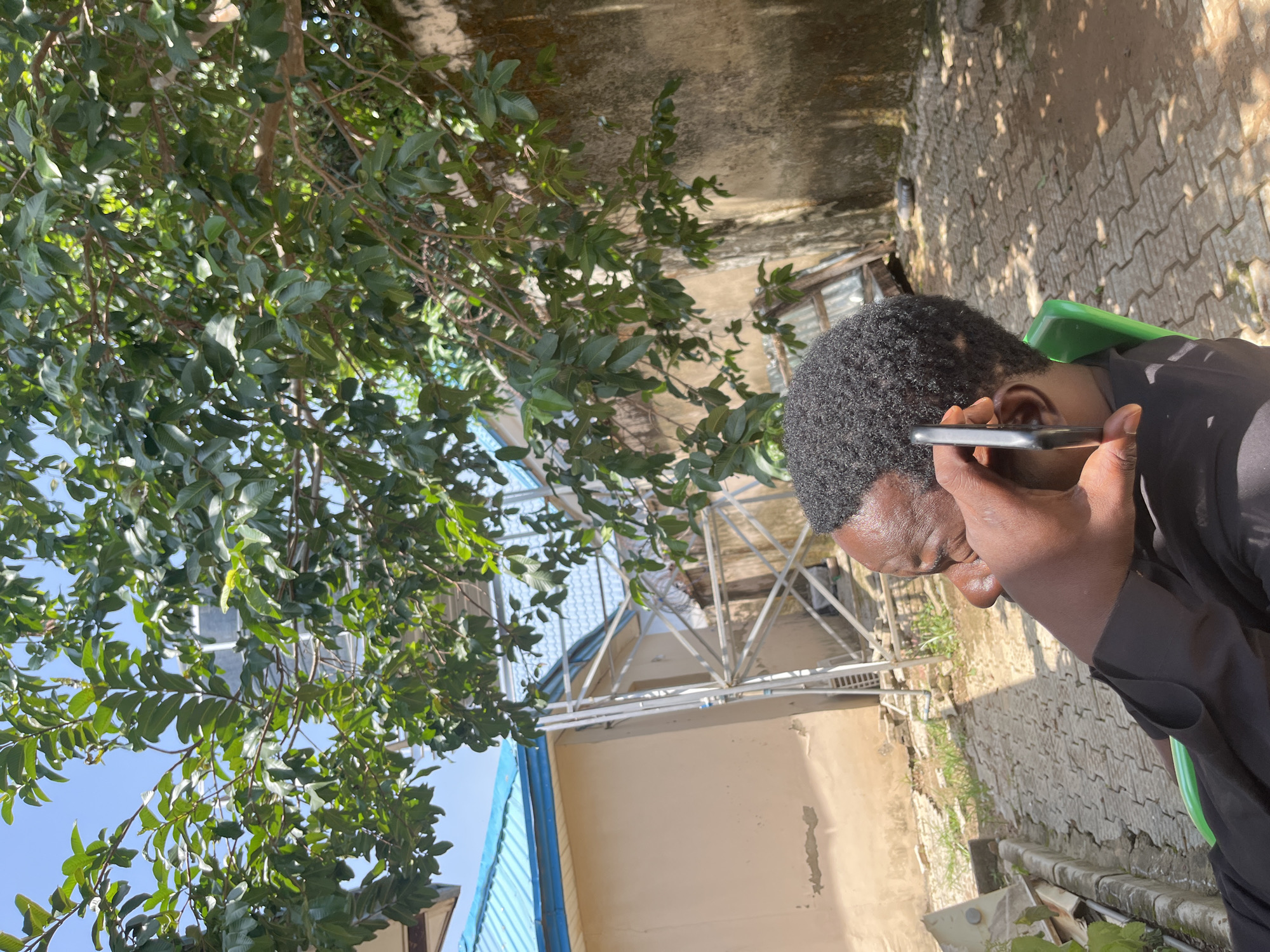 A person, with dark curly hair and dark skin, is holding a smartphone to their ear, appearing to be on a call. They are wearing a black shirt or tunic and are seated on a bright green plastic chair. The person is positioned in the middle of the frame, with their head tilted downwards.
The scene is outdoors, with a large leafy tree dominating the left side of the image, its branches extending across the upper portion of the frame. The background suggests an informal, possibly semi-rural or urban setting. There is a light-colored wall with visible signs of wear and tear behind the tree. Further back, there is a metal structure, possibly part of a building or scaffolding, with a blue-roofed structure visible. The ground is paved with interlocking bricks, and some patches of grass are growing between them.
The lighting suggests it is daytime, with shadows cast by the tree and structures indicating the sun is present. The overall atmosphere appears casual and everyday. No visible text is present in the image. The location context of Galadimawa Village, Nigeria, aligns with the visual cues of the outdoor setting, vegetation, and building styles.
