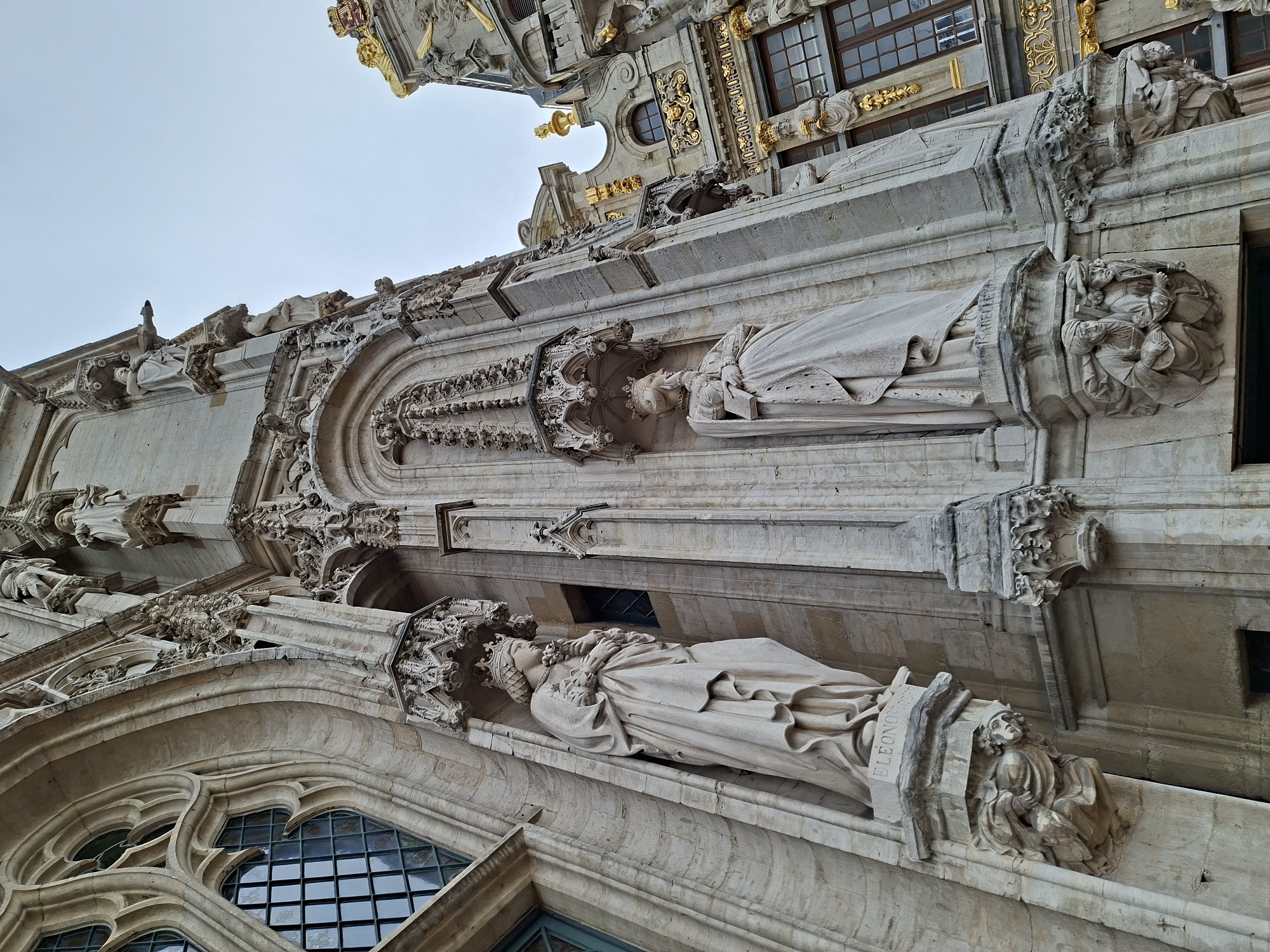 This is a low-angle, close-up, exterior shot of ornate, historical architecture in Brussels, Belgium, likely the Grand Place, given the style of buildings. The focus is on the detailed stonework of a building facade, featuring Gothic architectural elements. Two large, light-colored stone statues of robed figures wearing crowns are prominently displayed in niches. The statue on the left is labeled "ELEONORE" at its base, and the statue on the right is labeled "ISABELLE". These statues are supported by elaborate stone corbels carved with human figures in crouched positions. Above the niches, the building features intricate stone tracery and decorative architectural elements. To the right of the main facade, another building with gilded ornamentation and dark-paned windows is partially visible, suggesting a complex of grand, historic structures. The sky is overcast and a pale blue-grey, providing a neutral backdrop that emphasizes the detailed stonework of the architecture.