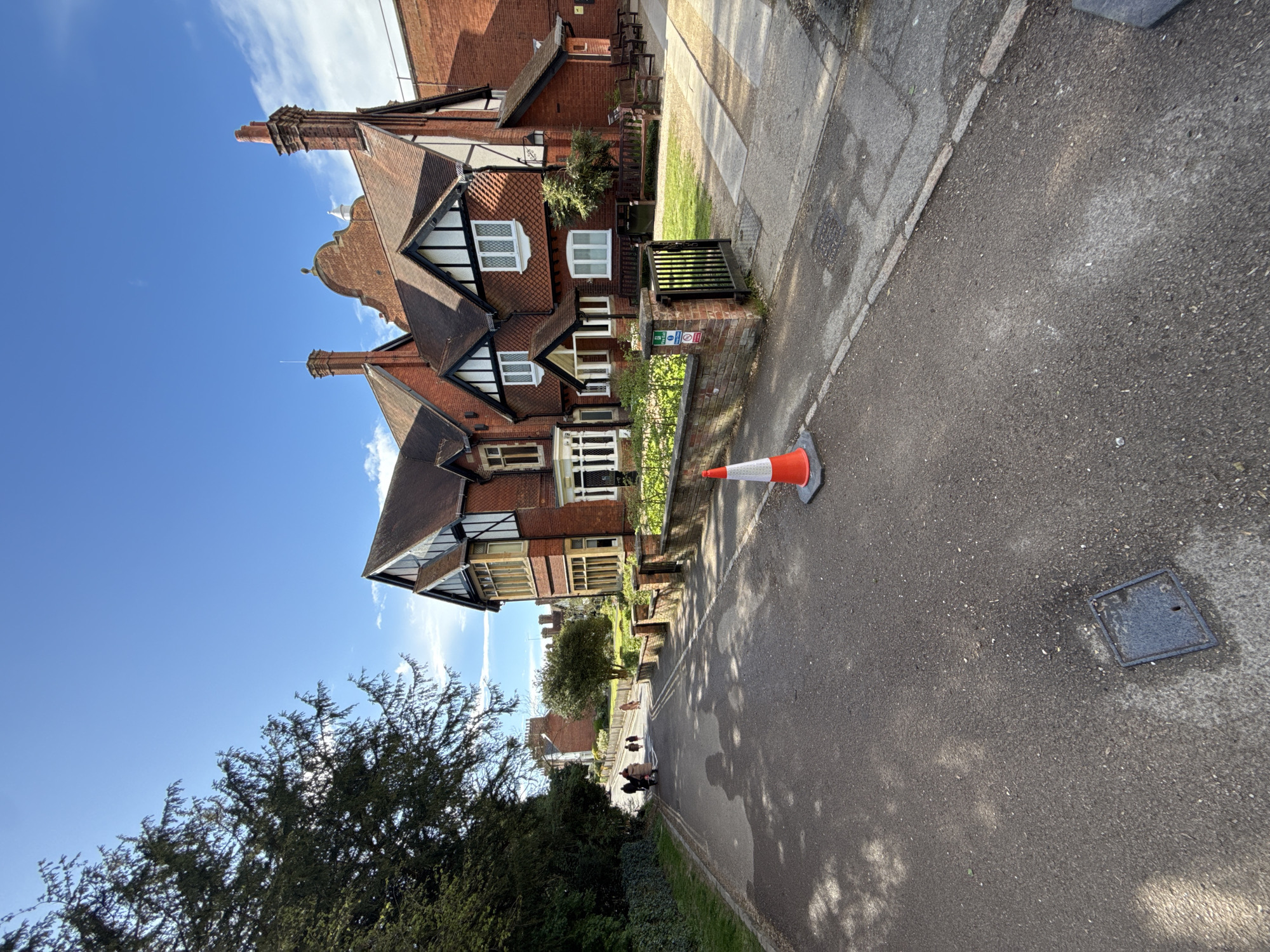 The image shows a residential street in Dacorum, United Kingdom, with a large brick building on the left side of the frame. This building features multiple gabled roofs, white-framed windows, and dark brown trim. A small garden area is visible in front of the building, with a short black metal fence separating it from the sidewalk. An orange and white traffic cone sits on the sidewalk near the building. The street is paved and appears to be in a residential area. A few people are seen walking in the distance. A tree is present in the foreground on the lower-left of the image. The sky is blue with some scattered clouds. A square manhole cover is visible on the street.