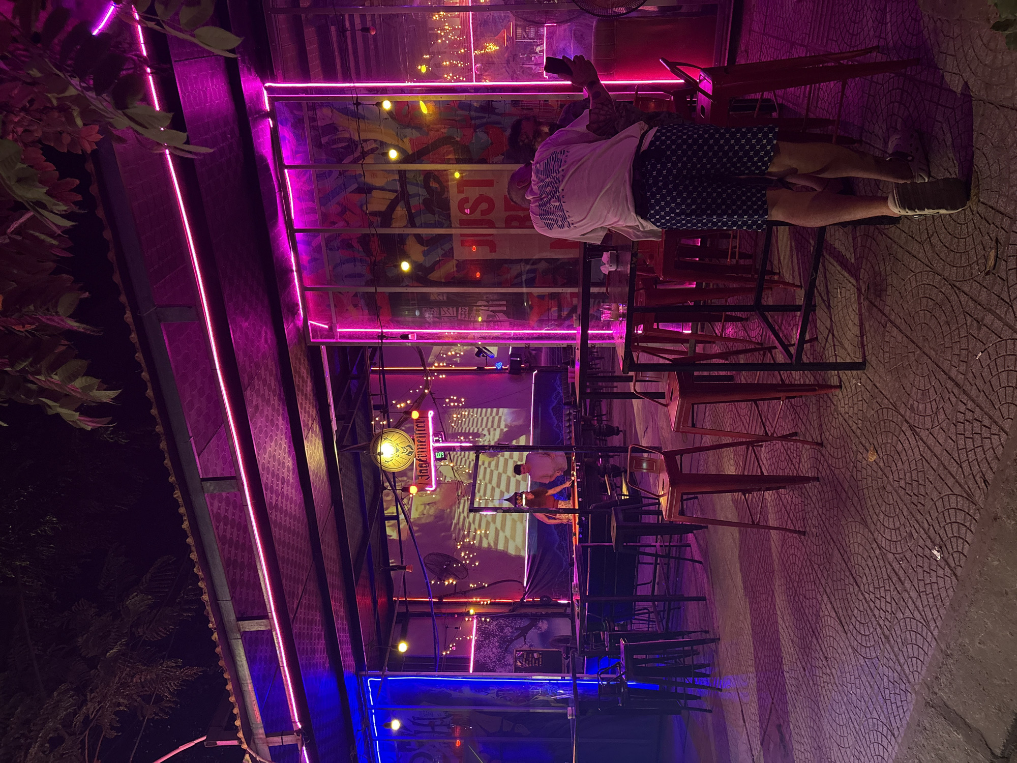 A nighttime scene in Đà Nẵng, Vietnam, depicts an outdoor bar area. A partial view of a man in a white t-shirt and dark patterned shorts stands at a high-top table, holding a smartphone. Several empty high-top tables with orange bar stools are present. The bar is partially covered with an awning, decorated with pink neon lights, and a lower section illuminated by blue lights. A wall behind the bar has a mural or screen displaying images and words, with beer branding visible (Jägermeister). The ground is paved with a patterned surface. Trees are partially visible on the left side of the frame.