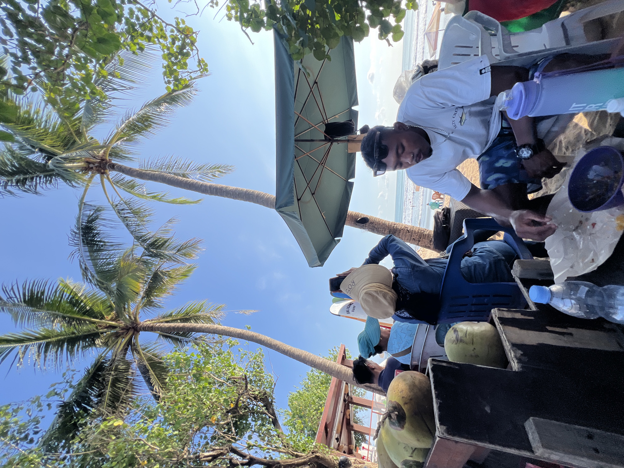 The image depicts a beach scene in Kuta, Indonesia, characterized by palm trees, a clear blue sky, and the ocean in the background. In the foreground, several green coconuts are arranged on a wooden stand. To the right, a man wearing sunglasses and a white t-shirt with a graphic print sits on a white plastic chair, holding a paper plate with food. A purple and blue water bottle is visible next to him. In the middle ground, a woman wearing a straw hat is taking a photo with her phone, while other individuals are seated nearby. Two surfboards are leaning against a railing. A green umbrella provides shade for a seating area. The beach appears to be a popular spot with several people and umbrellas visible in the distance.