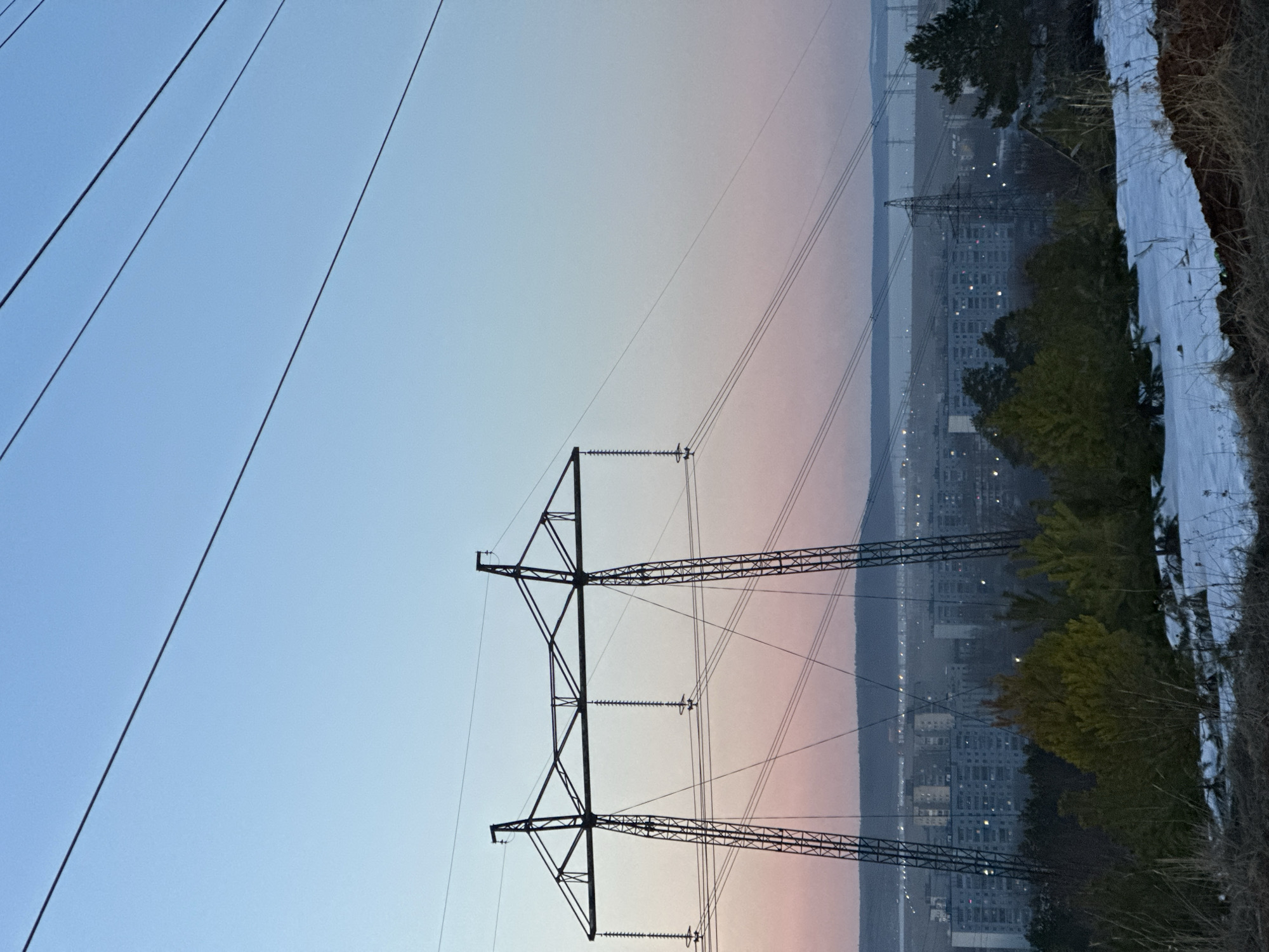 The image shows a view in Chaikovsky, Russia, featuring power lines and a transmission tower against a backdrop of buildings and a skyline. The sky transitions from blue to a faint pink hue. Trees and snowy terrain are visible in the foreground on the right. The power lines stretch across the sky, and the tower stands prominently in the middle of the image.