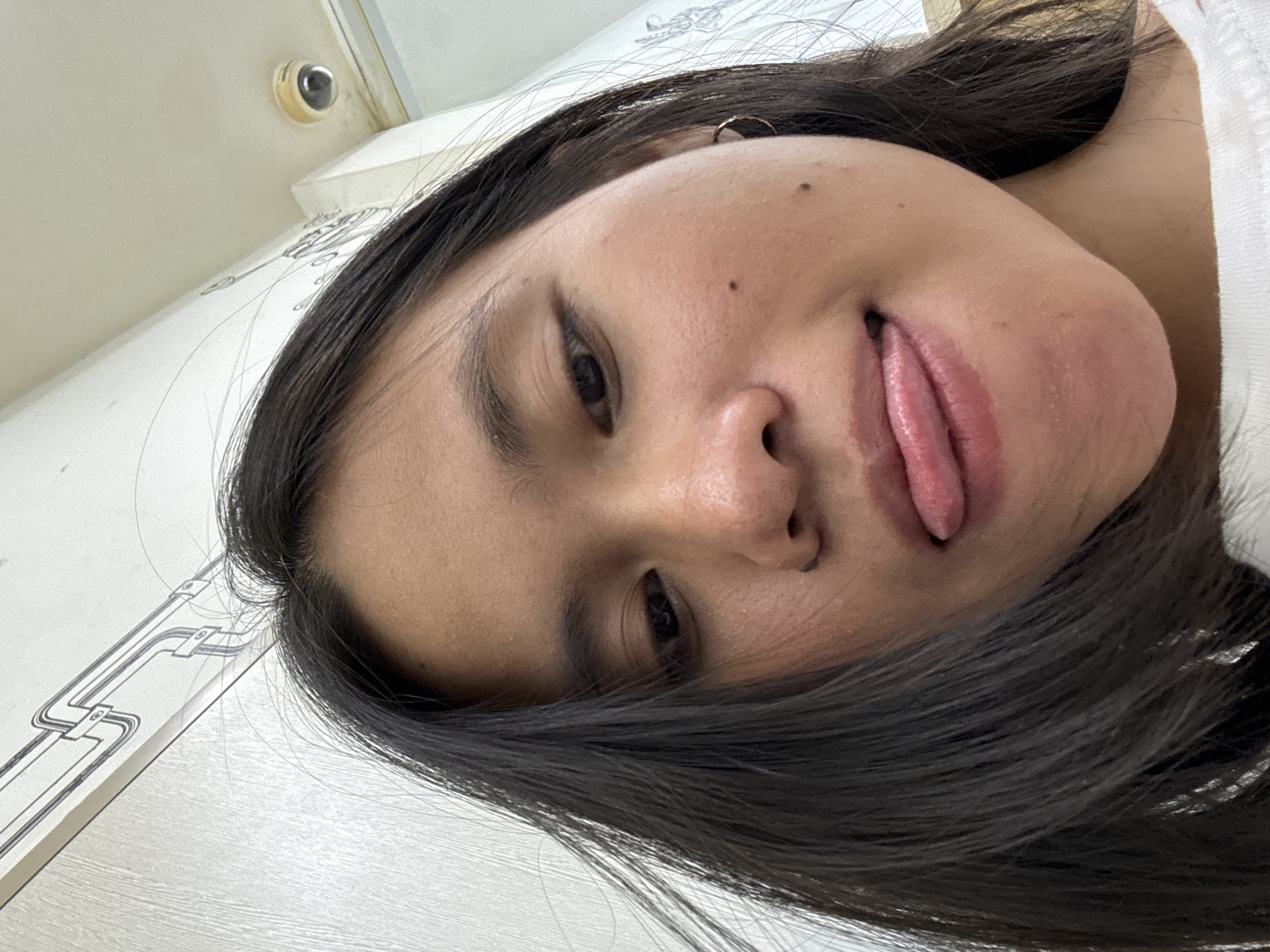 A close-up, low-angle selfie shows the face of a young woman with long, dark hair. She is sticking her tongue out, and her mouth is slightly open. She has a small mole on her cheek and a gold hoop earring in her visible ear. In the background, a white wall with a linear, patterned design is visible, along with a white ceiling and a small surveillance camera. The scene is indoors, likely in an urban residential or commercial setting. It is not possible to determine the specific location in East Jakarta, Indonesia, from the provided image.