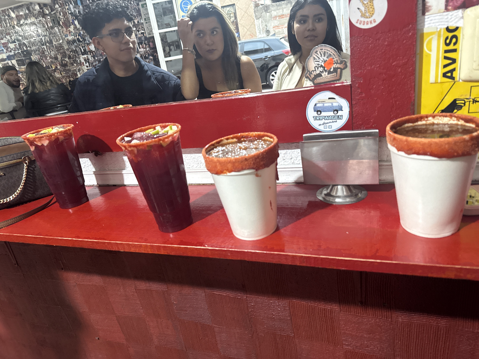 The scene depicts a food stall in San Andrés Cholula, Mexico, where four drinks stand on a red counter. From left to right: a dark red drink in a clear plastic cup, a similar but slightly lighter drink with diced fruit on top, a brownish drink in a white cup, and a light-colored drink, also in a white cup. All cups have a bright red rim. Behind the counter are three people. A young man with glasses is on the left, and two women stand to his right. The stall's backdrop is cluttered with stickers, signs, and an accumulation of photos. A car is visible through the stall, indicating an outdoor or semi-outdoor location.