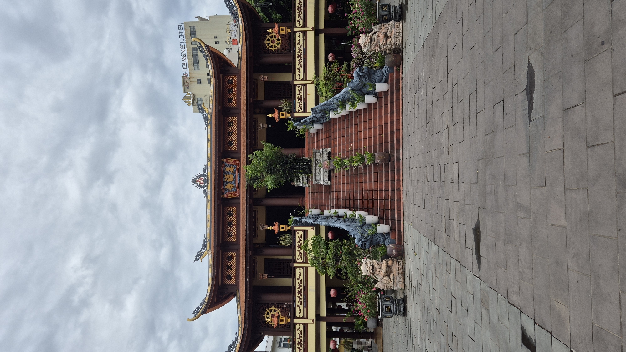 The image shows the exterior of a temple, likely a Buddhist temple, with traditional East Asian architectural elements. The temple has a multi-tiered, curved roof with ornate detailing and a rich brown and gold color scheme. A wide, red staircase leads up to the entrance of the temple, flanked by stone dragon sculptures and potted plants. The courtyard in front of the temple is paved with large, flat grey stones, some of which appear damp. To the right and in the background, a modern hotel building with the sign "DIAMOND HOTEL" is visible, indicating an urban setting in Đà Nẵng, Vietnam, where this temple is situated. The sky is overcast with heavy grey clouds. There are no people visible in the image.