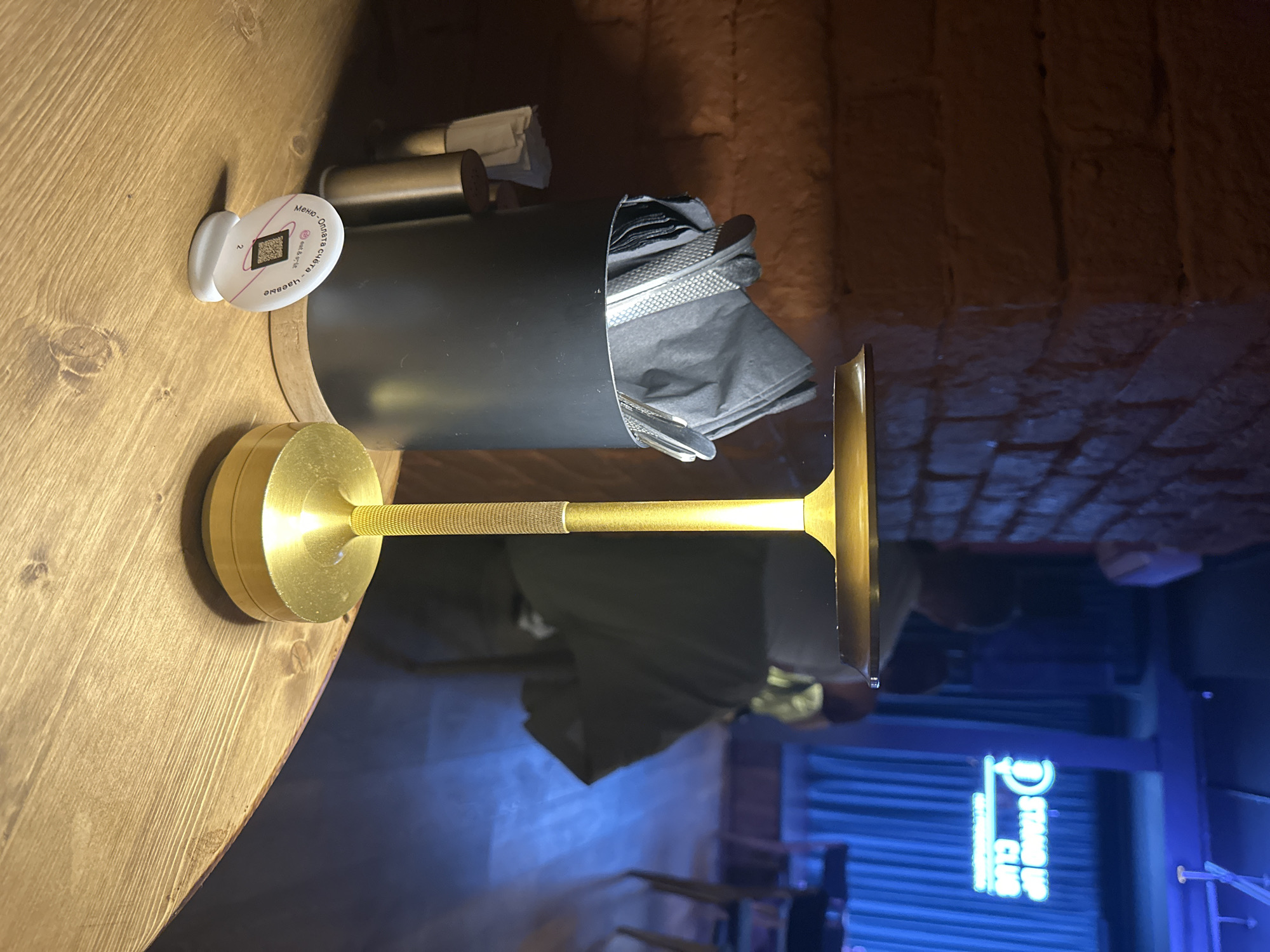 The image captures a dimly lit interior setting in what appears to be a bar or restaurant. On a wooden table, there is a golden-colored metal napkin holder filled with black napkins. A white disc with a QR code and text in Russian is affixed to the table next to the napkin holder. The text on the disc appears to say "Menu and tips" (translated from Russian). In the background, there is a brick wall. Through a window, a blurred neon sign is visible, which reads "STAND UP CLUB". Based on this sign, the location is likely in Saint Petersburg, Russia, as this is the only city where "Stand Up Club" is available. No people are visible in the frame. The scene is generally suggestive of a place where patrons might gather for entertainment and refreshments.
