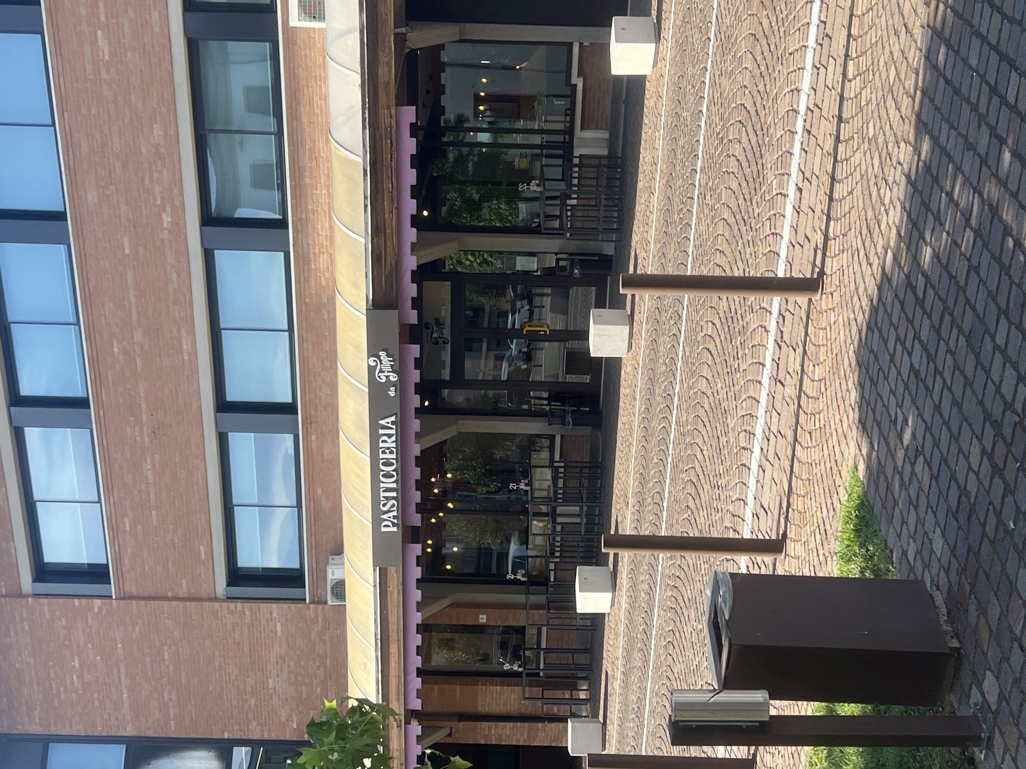 This is a vertical, eye-level shot of a building facade with a pastry shop. The building is made of reddish-brown brick with large, dark-framed windows. The shop, "PASTICCERIA da Filippo," has a yellow awning and a glass storefront. The entrance is marked by two sets of glass doors. Inside, the shop is lit with warm, ambient light, and trees are visible through the glass, suggesting an outdoor seating area or a view of greenery.
The scene is outdoors on a bright, sunny day, indicated by the strong shadows cast on the cobblestone pavement and the bright lighting. The pavement is laid in a diamond pattern, typical of many Italian streets and piazzas. There are several brown metal bollards and a dark brown trash receptacle visible along the walkway. The overall atmosphere is that of a peaceful, ordinary day in a European town.
There is no visible activity or event taking place; the scene appears static, focusing on the architecture and the shop's exterior. The time of day is likely midday, given the angle of the shadows. The location is identified as Campodarsego, Italy, which is further supported by the architectural style and paving. The text "PASTICCERIA da Filippo" is clearly visible on a dark sign above the shop's entrance.