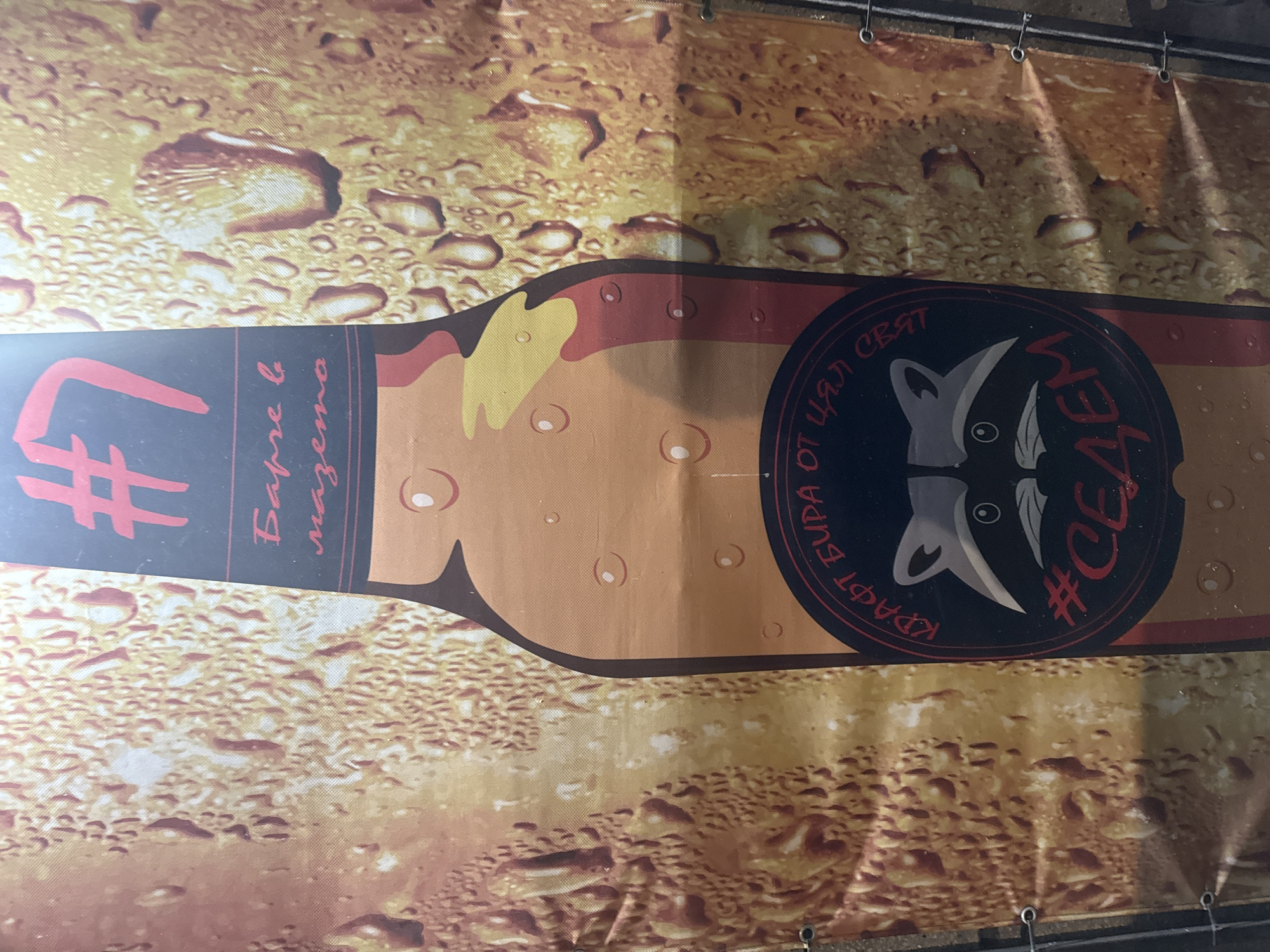 A banner depicting a beer bottle on a background that simulates condensation on a beer glass is visible. The bottle's neck is dark with a red hashtag symbol and the number "7". Beside that, the Cyrillic inscription reads "Баре в мазета". The body of the bottle is light brown and suggests a foam head, with a circular black logo placed on it. Inside the logo, a stylized image of a raccoon's head appears. Red Cyrillic text around the logo reads "КРАФТ БИРА ОТ ЦЯЛ СВЯТ" and "#СЕЯЕМ". The banner appears to be hanging with metal grommets, although the background is unclear. The city of Varna, Bulgaria is not discernable from the image.