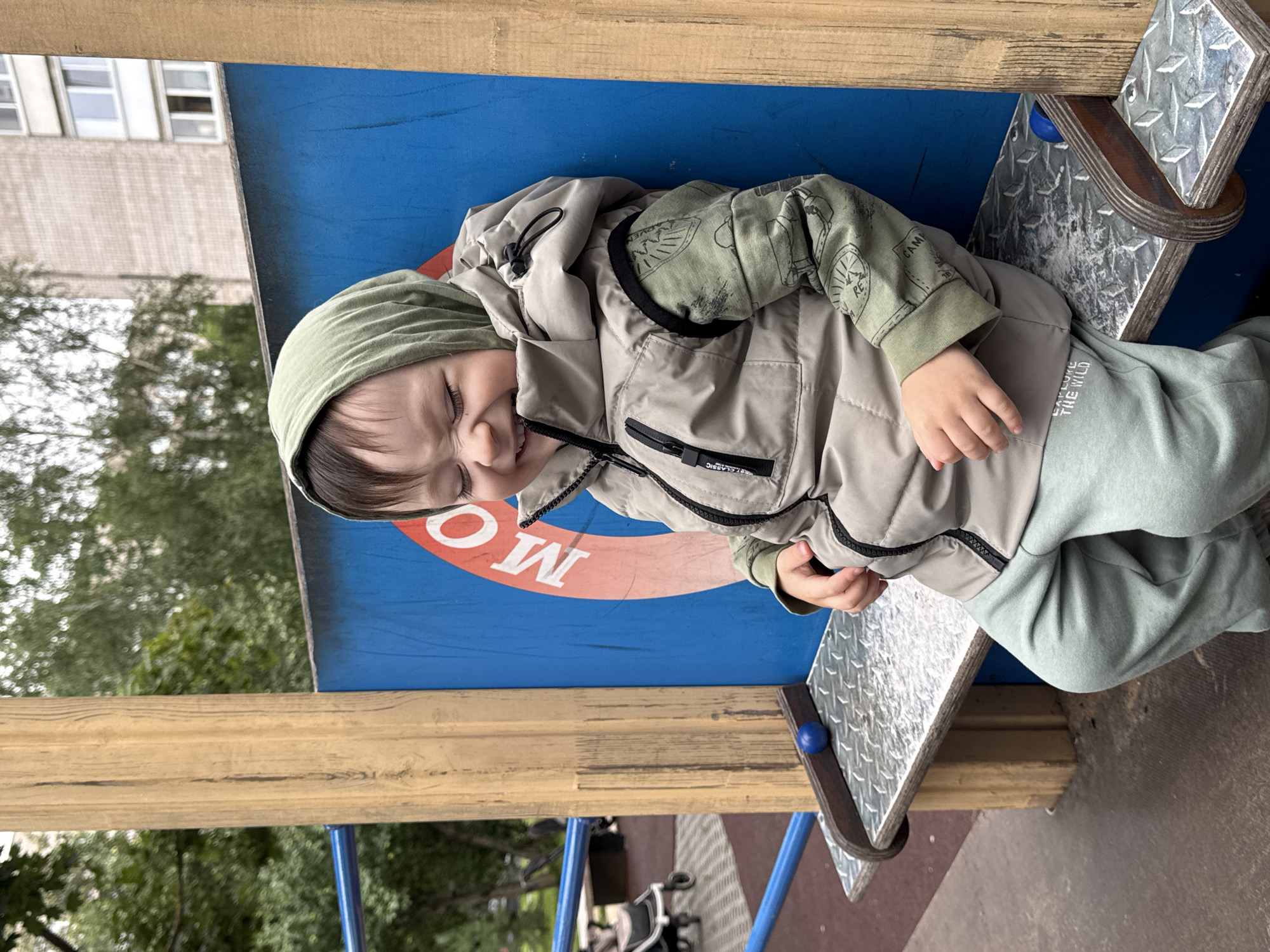 A young child with brown hair, wearing a green hood, a light brown vest over a green long-sleeved shirt with a print, and light green sweatpants, is sitting on a metal bench. The child is smiling with their eyes closed and head tilted back. The bench is part of a playground structure with a blue panel behind it, featuring a red circle with white lettering. To the left of the bench, there is a wooden post. In the background, trees and a multi-story building are visible. The ground appears to be paved. The scene is outdoors in an urban environment, likely a playground in Izhevsk, Russia.