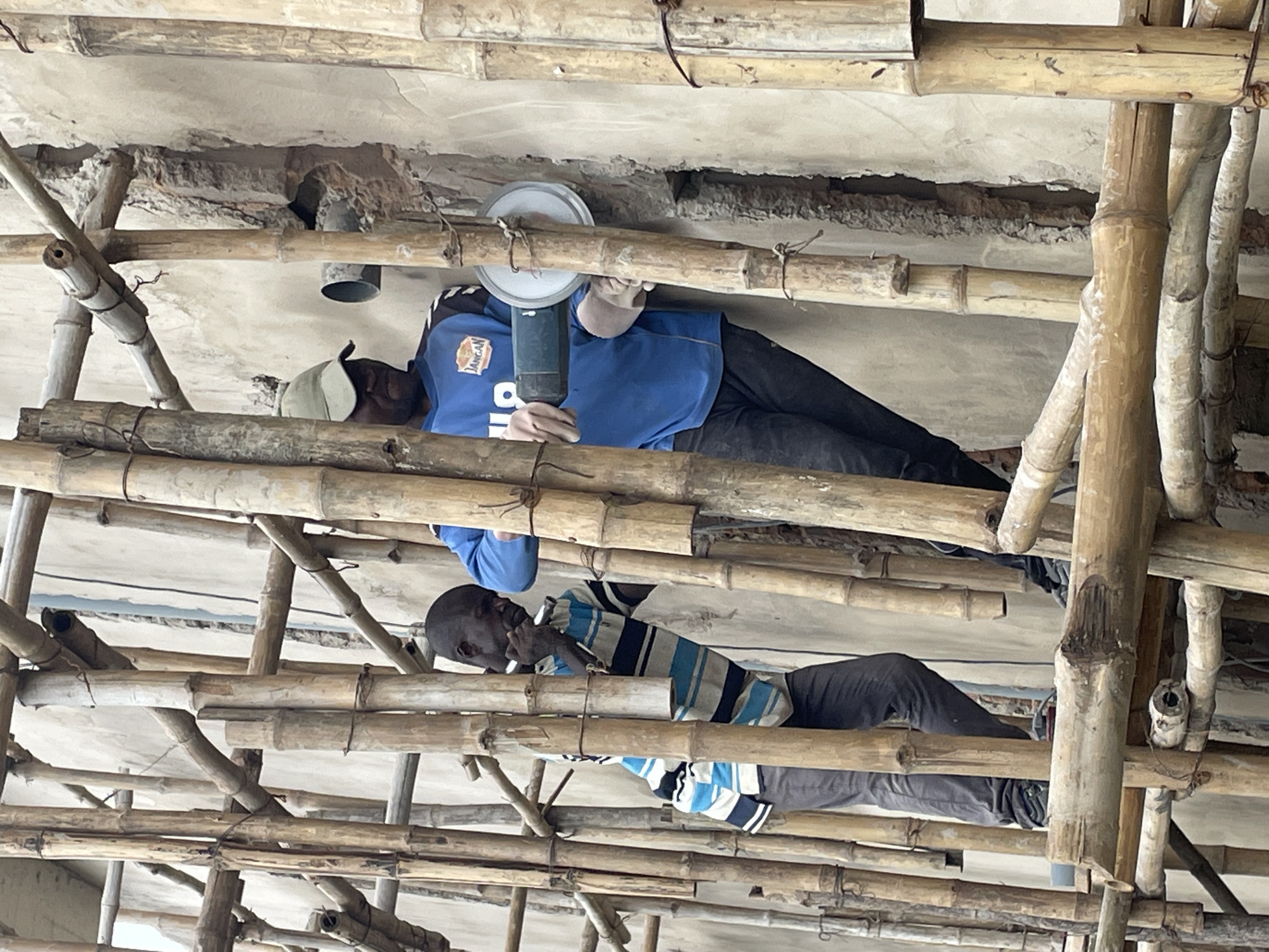In Okinano, Nigeria, two men are working on scaffolding constructed from bamboo poles. The man in the foreground, wearing a blue shirt with a white logo and a green cap, is using a grinder to cut into a concrete wall. He is wearing light-colored gloves. To his left, another man, wearing a horizontally striped blue and white shirt and dark pants, is also working on the wall, holding a tool. The concrete wall appears damaged and broken in places. The scaffolding extends upwards and across the frame, obscuring parts of the wall and the surrounding environment.