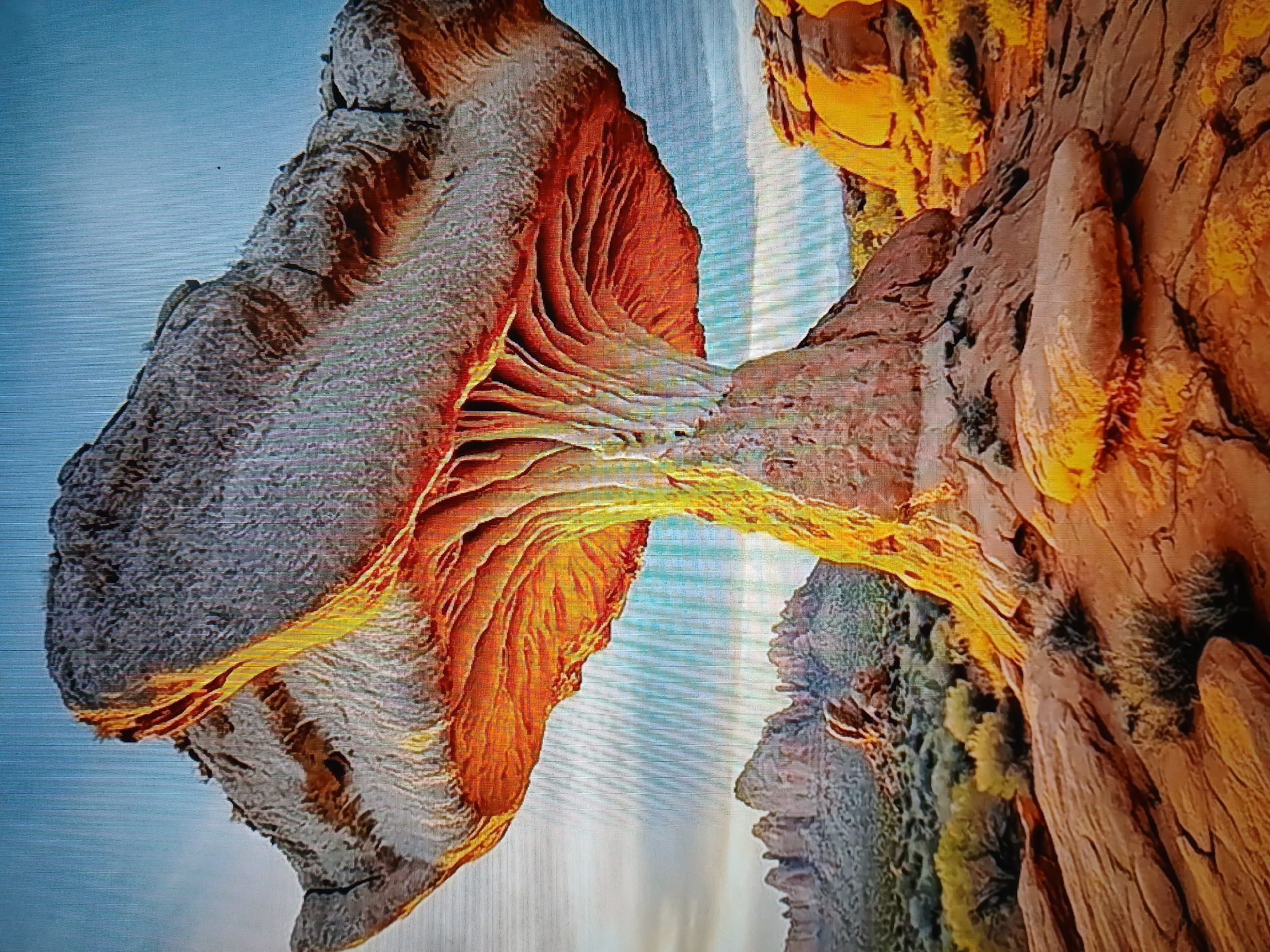 The image shows a large rock formation in a desert landscape, resembling a mushroom. The cap of the "mushroom" is a broad, flat stone, while the stem is composed of thinner, striated rock formations that create an umbrella-like effect. The underside of the cap is illuminated with a warm, orange glow, suggesting either natural sunlight or artificial lighting. The surrounding environment consists of arid terrain with sparse vegetation and distant rock formations, indicative of a desert or canyon region. The sky is a clear blue with some faint cloud cover. The perspective is from a low angle, emphasizing the scale and dramatic appearance of the rock formation. There are no people or animals visible in the image. The overall impression is one of natural geological artistry in a remote, undeveloped area.