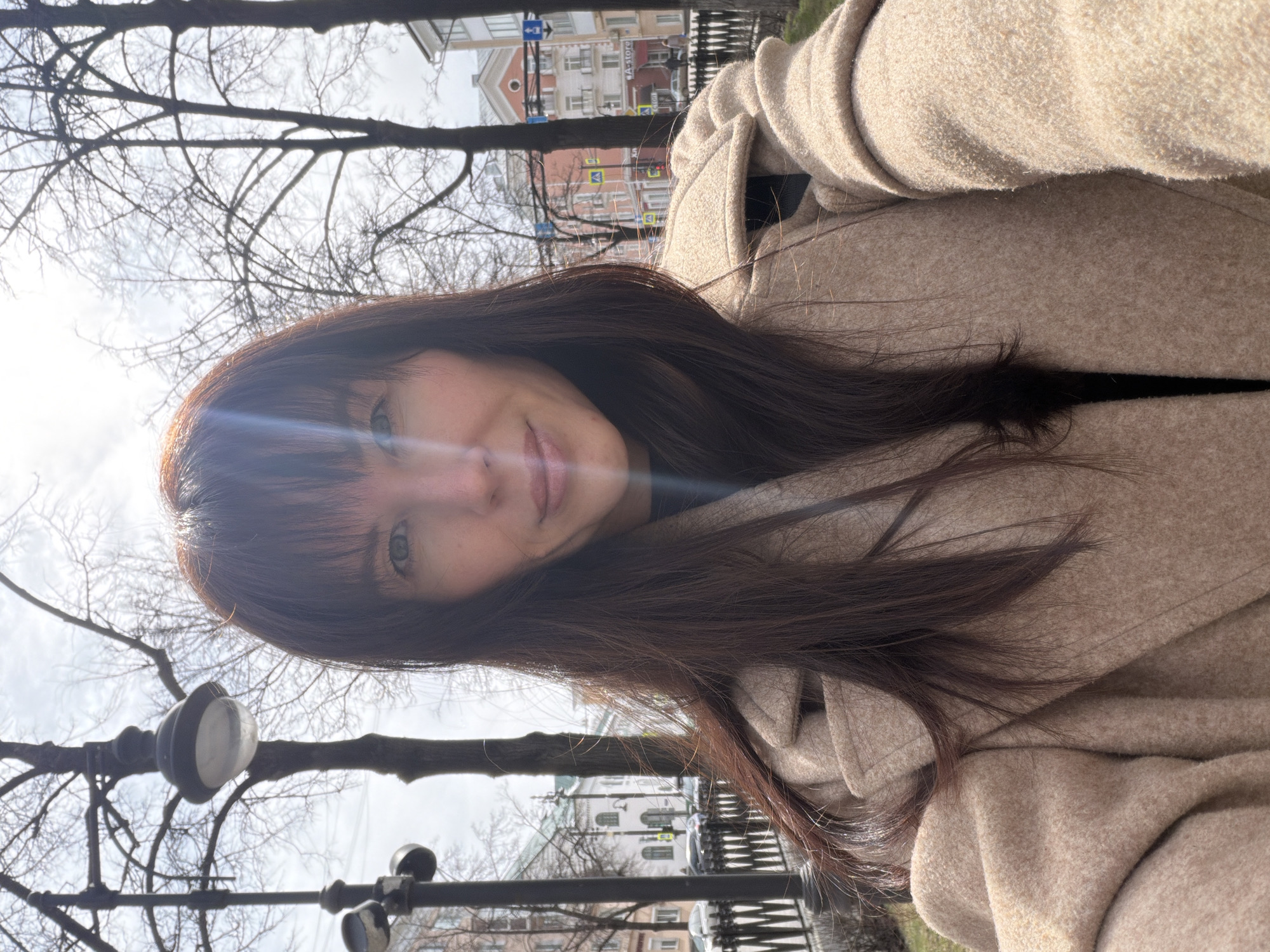 A woman with long, brown hair and bangs, wearing a tan coat, is taking a selfie outdoors. The background reveals a cityscape, suggesting an urban setting. Bare trees line the street behind her, and street lights are visible. The style of architecture in the distance indicates the photograph was taken in Perm, Russia.