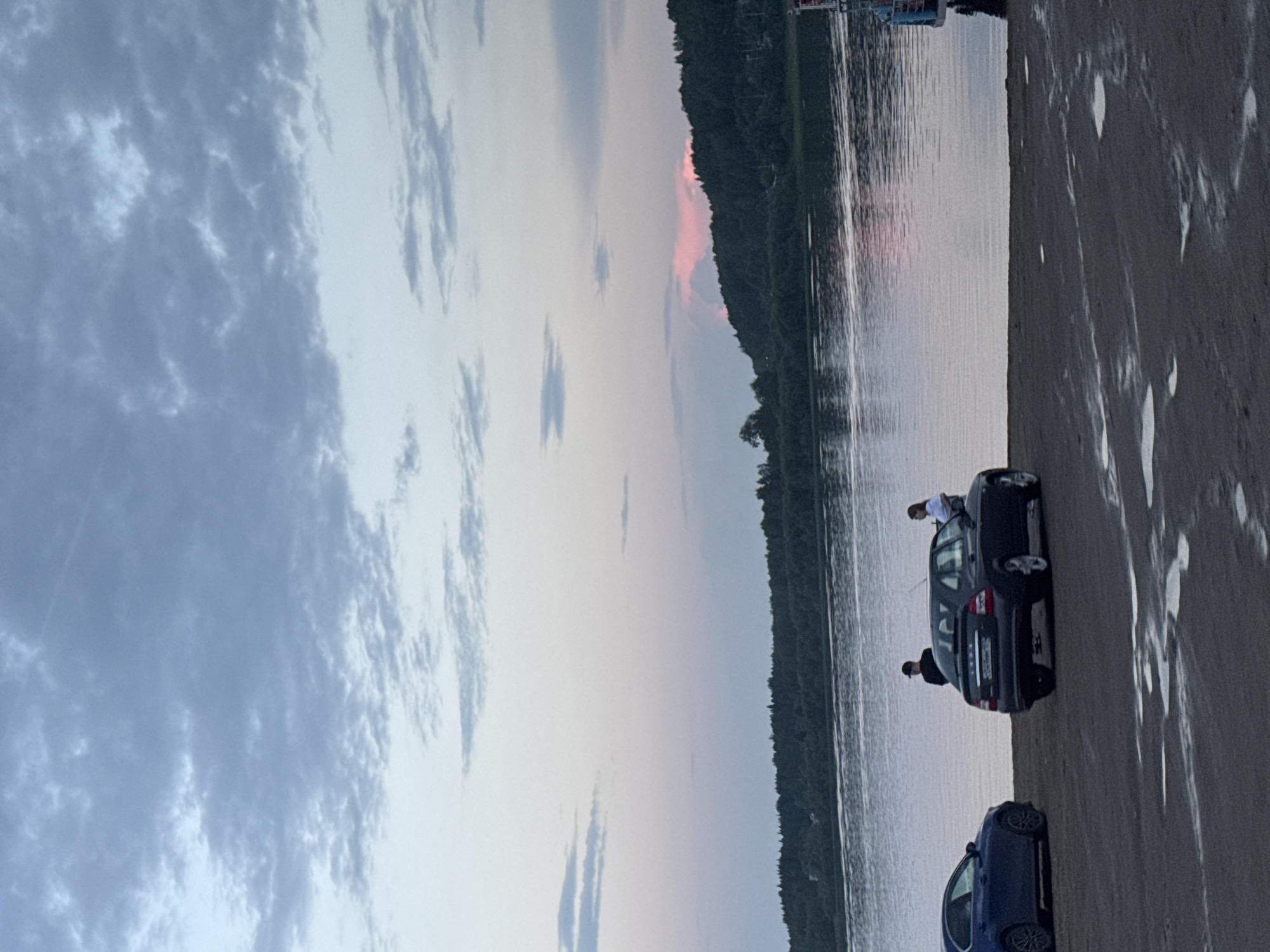 The image depicts a waterfront scene at dusk in Tyulkino, Russia. The sky is filled with a gradient of pale blue and soft pink clouds, with a hint of pink light reflecting on the water near the horizon. A body of water stretches across the middle ground, meeting a densely forested shoreline on the opposite bank. In the foreground, a dark sandy area shows patches of moisture.
On the sandy bank, two cars are parked. A dark-colored sedan is positioned closer to the viewer, facing slightly to the right. A lighter-colored sedan is parked to its left, facing the water. Two individuals are standing near the dark sedan. One person, wearing a dark shirt and a cap, stands behind the car. Another person, in a light-colored shirt, is leaning against the trunk of the dark sedan. The scene suggests a moment of leisure by the water.
