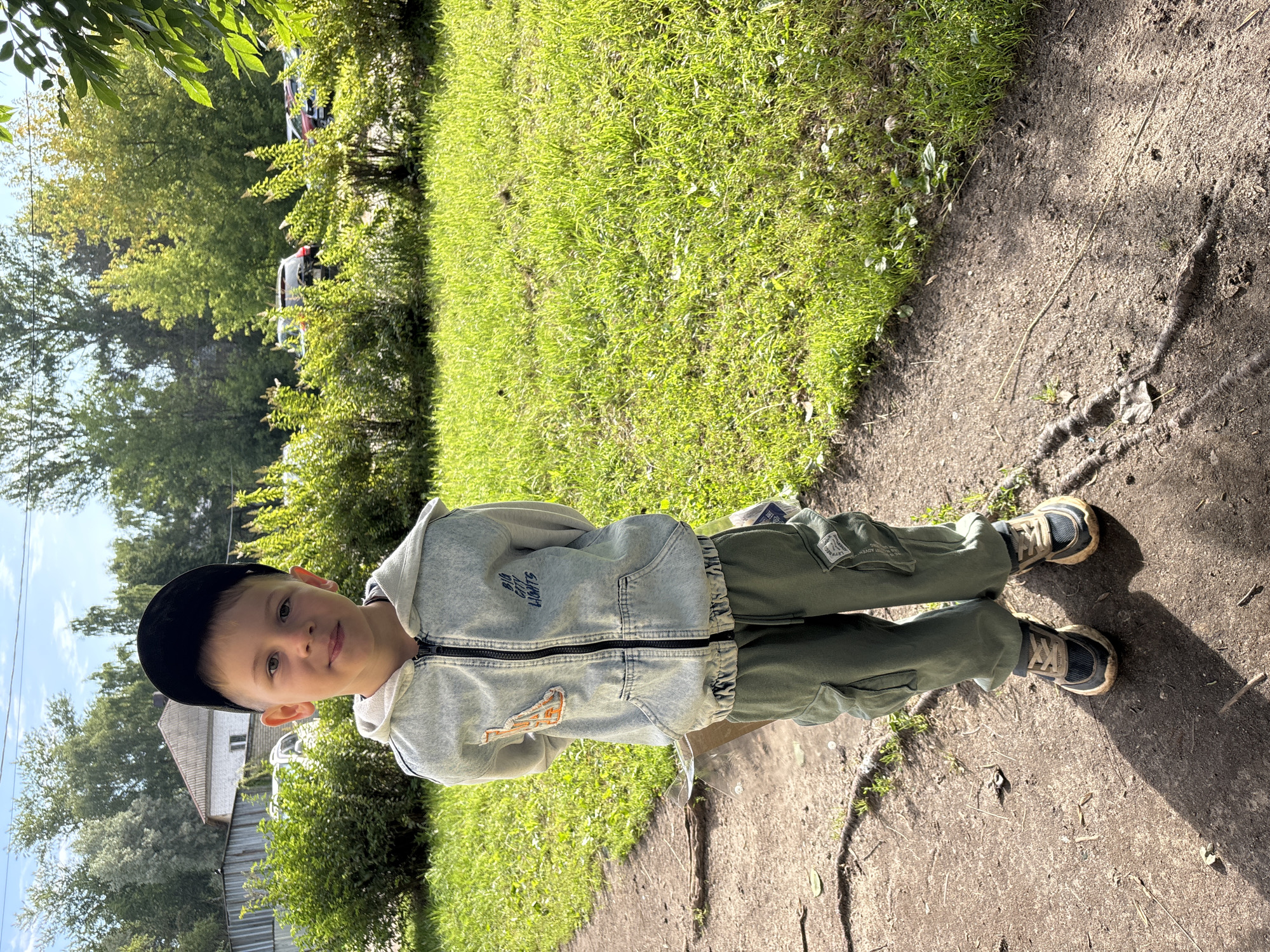 A young boy wearing a black baseball cap and a light blue denim-look hoodie stands outdoors on a paved path next to a grassy area. He is facing the camera with his hands behind his back. The boy is wearing green cargo pants and dark-colored sneakers with tan laces. In the background, there are green bushes, trees, and parked cars. A white building is partially visible behind the bushes. The scene appears to be in a residential area. The image was taken in the city of Rusanovo, Russia.