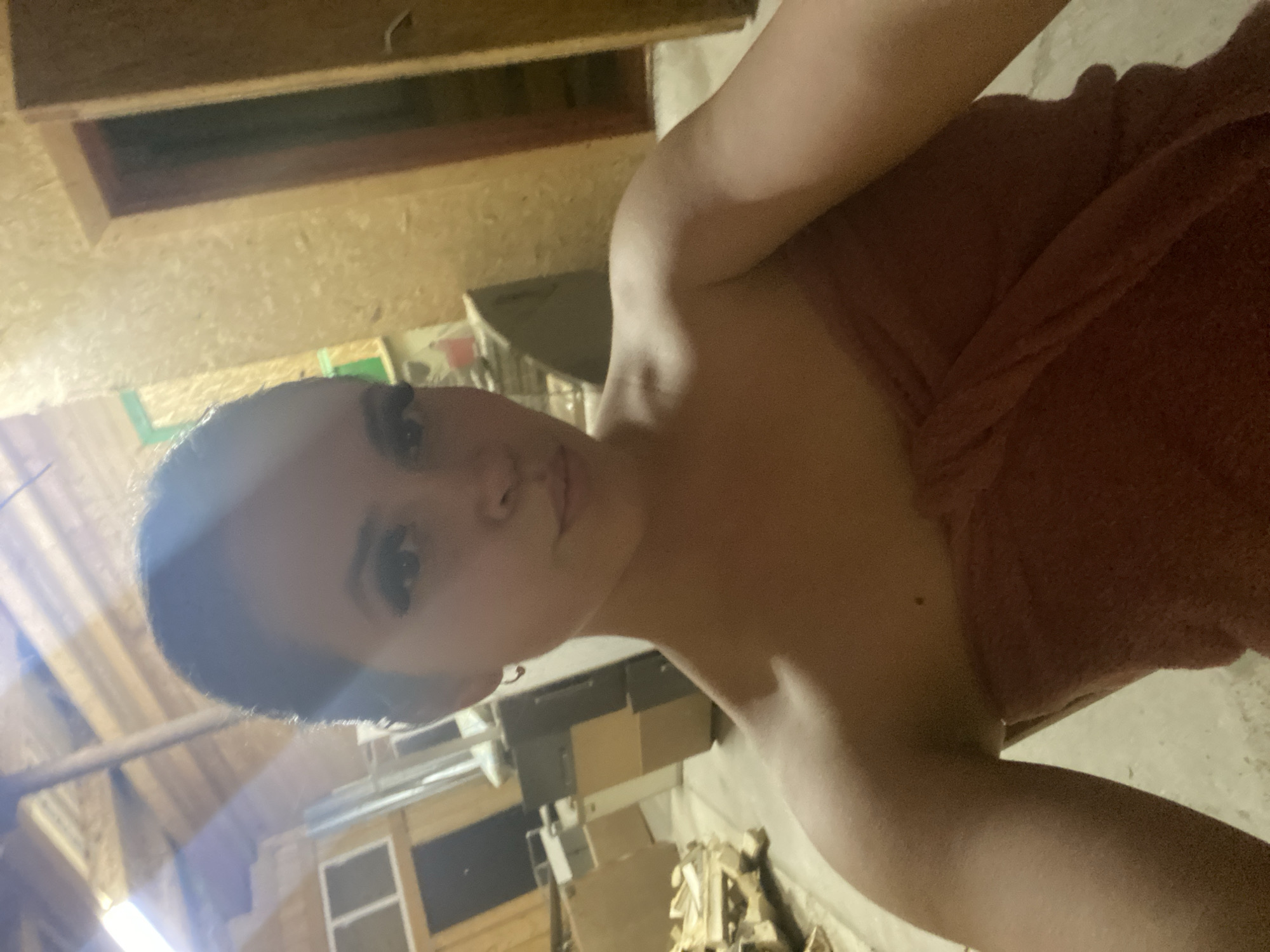 A selfie-style photograph shows a woman from a low angle, looking directly at the camera. She appears to be wrapped in a reddish-brown towel. The background suggests an interior space, possibly a basement or storage area, with plywood walls and ceiling rafters visible. Other objects such as boxes and equipment are also present. The location in Zhigalki, Russia is not identifiable.