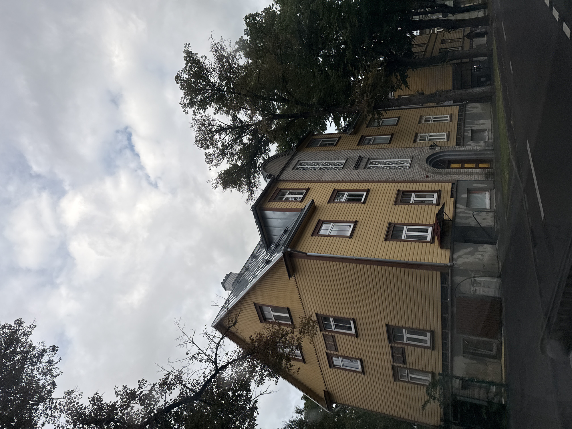 The image depicts a yellow wooden building in Tallinn, Estonia, on a cloudy day. The building has multiple stories, with a pitched metal roof and several windows. The facade is clad in horizontal wooden planks, with a section of gray brickwork around the entrance. Trees with dark green foliage partially obscure the building from the left and right sides. The building is situated on a street with a paved road and visible white markings. The overall atmosphere is overcast and somber.
