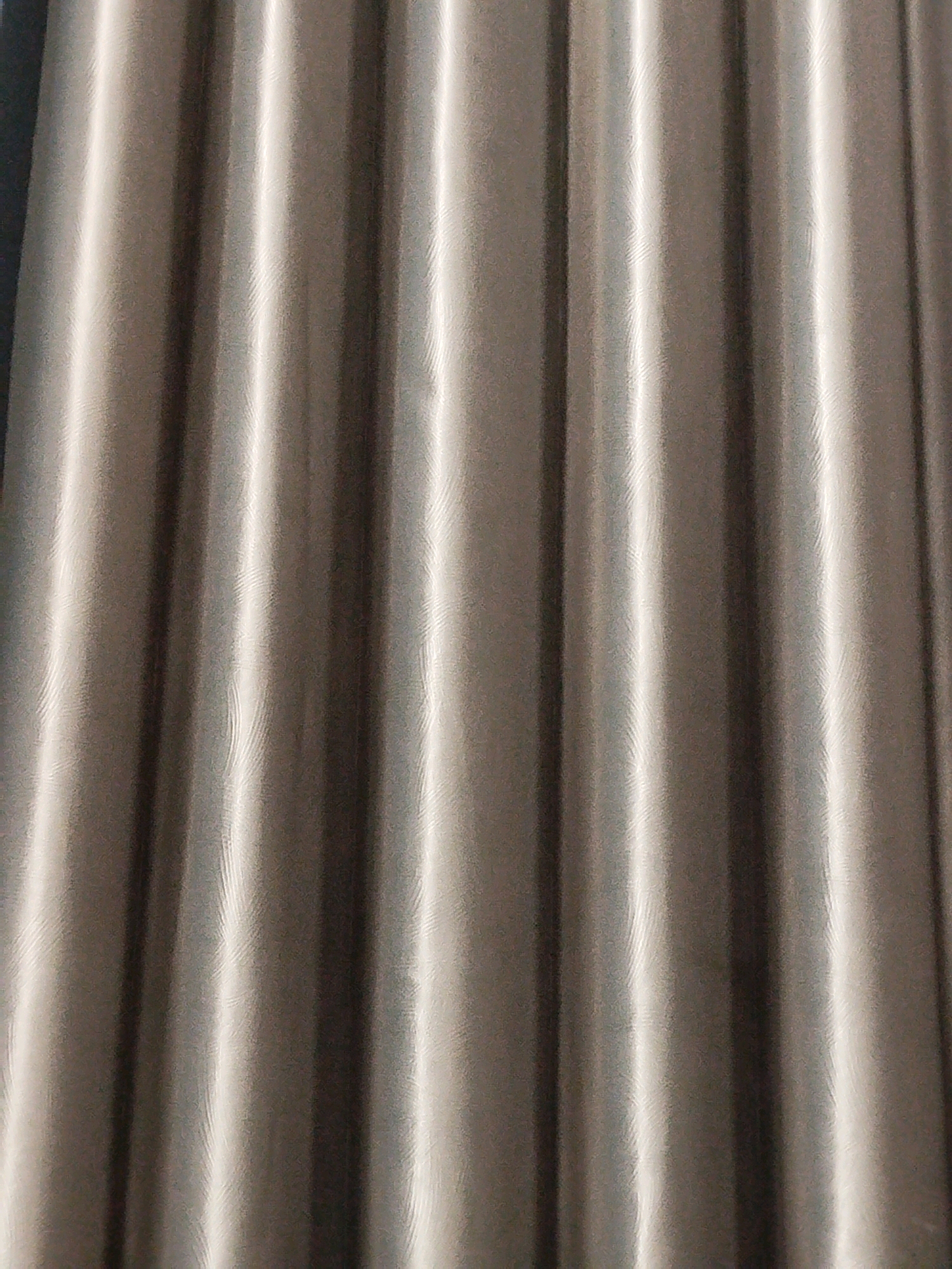 The image shows a close-up, abstract view of a corrugated metal surface. The texture is characterized by vertical ridges and valleys, with strong highlights and shadows that create a sense of depth and metallic sheen. The color palette is dominated by shades of gray and silver, suggesting a brushed or polished metal finish. The repetitive pattern of the corrugation fills the entire frame, offering no discernible context of its surroundings, such as a specific location in Mojoagung, Indonesia, or any people, objects, actions, interactions, or environmental details.