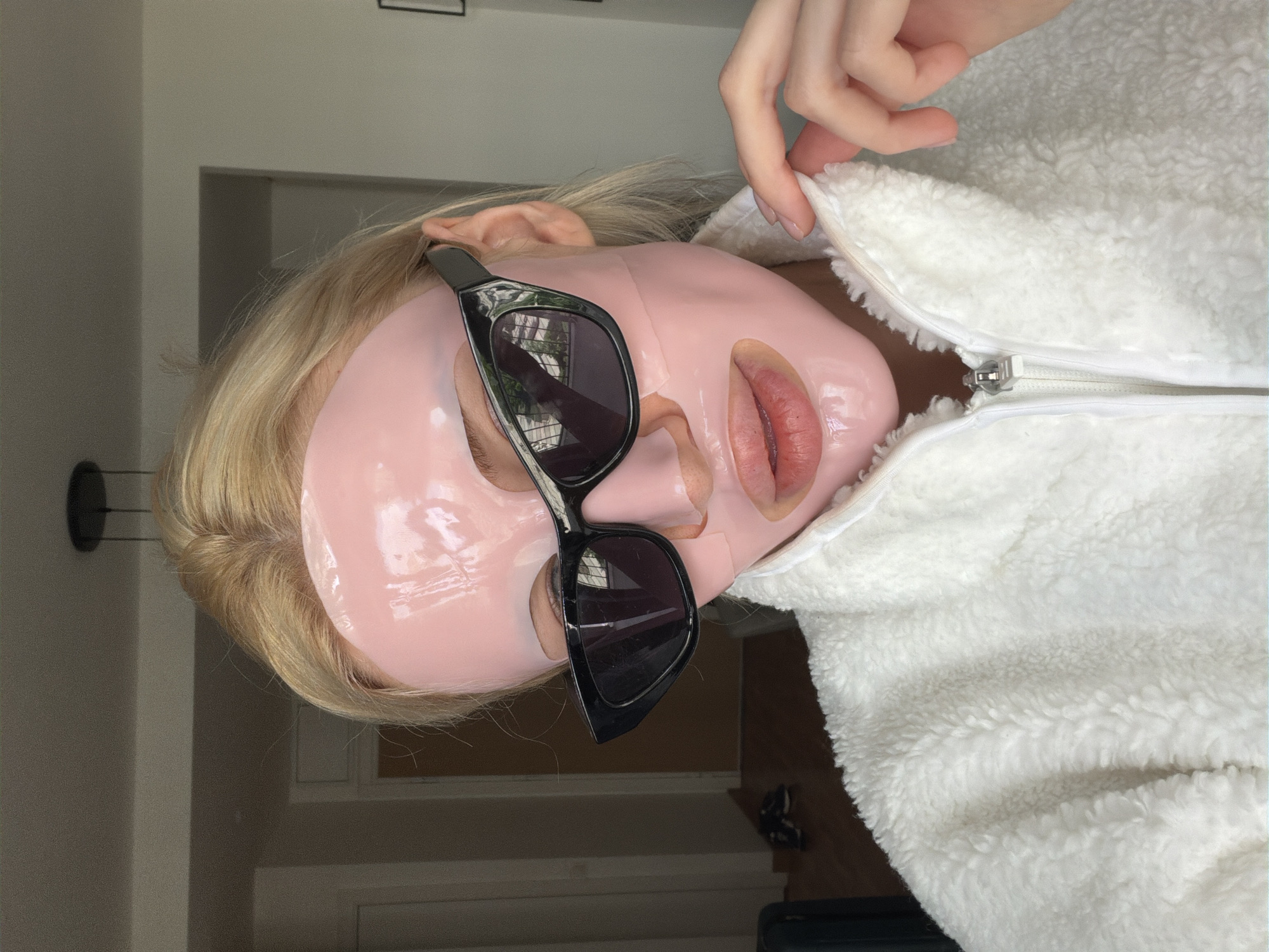 A person with blonde hair is wearing a pink facial mask and black sunglasses. They are wearing a white, fuzzy jacket with a zipper. The background shows a doorway and a wall. The location cannot be determined from the image alone.