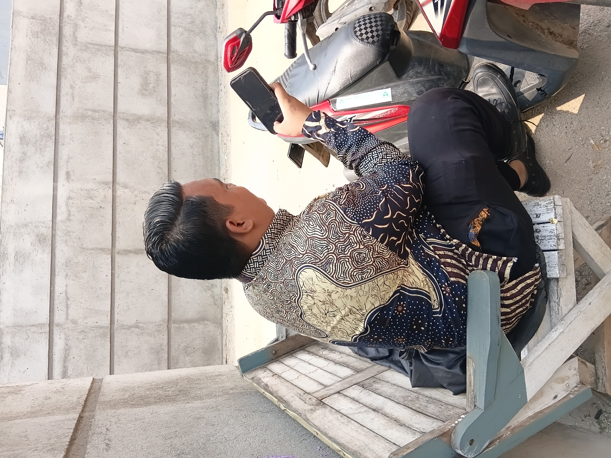 A man wearing a batik shirt and black pants sits on a wooden bench next to a red motorcycle in what appears to be an outdoor urban setting in Bekasi, Indonesia. He holds a black smartphone in his right hand and is looking at it. The background consists of a textured concrete wall. The bench is weathered and appears to be made of light-colored wood with blue accents. The motorcycle is parked to the man's right, partially visible. The ground is covered with sand and small stones. The lighting suggests it is daytime with a strong light source, casting shadows.