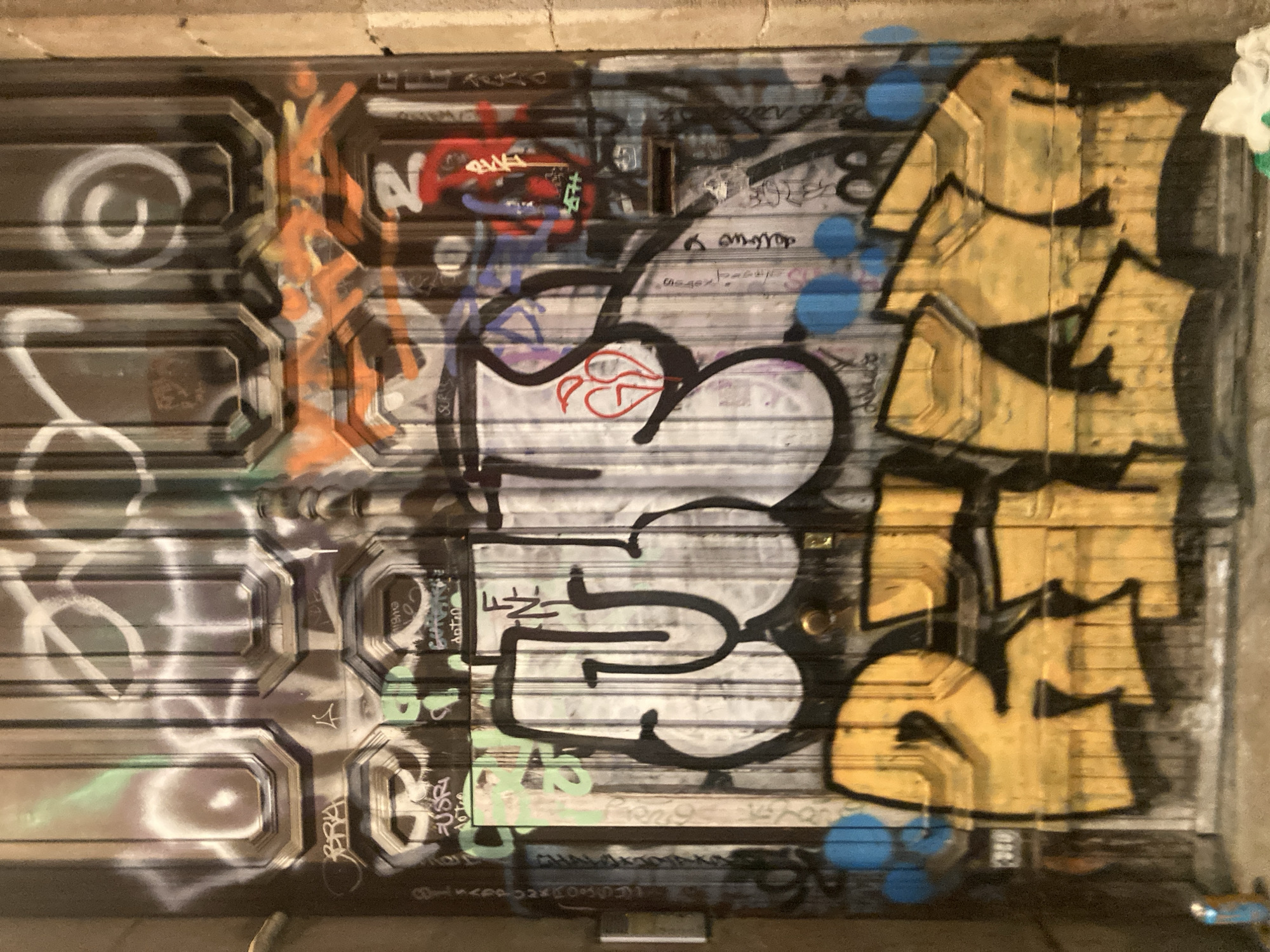 The image shows a metal door covered in graffiti in Barcelona, Spain. The door is dark brown with octagonal panels. The graffiti is layered and varied, including large, bold yellow letters across the bottom, black outlines filled with white and grey on the upper half, and smaller tags and scribbles in various colors including orange, red, and green. There are also blue circles painted on the door. The overall impression is of urban decay and street art.