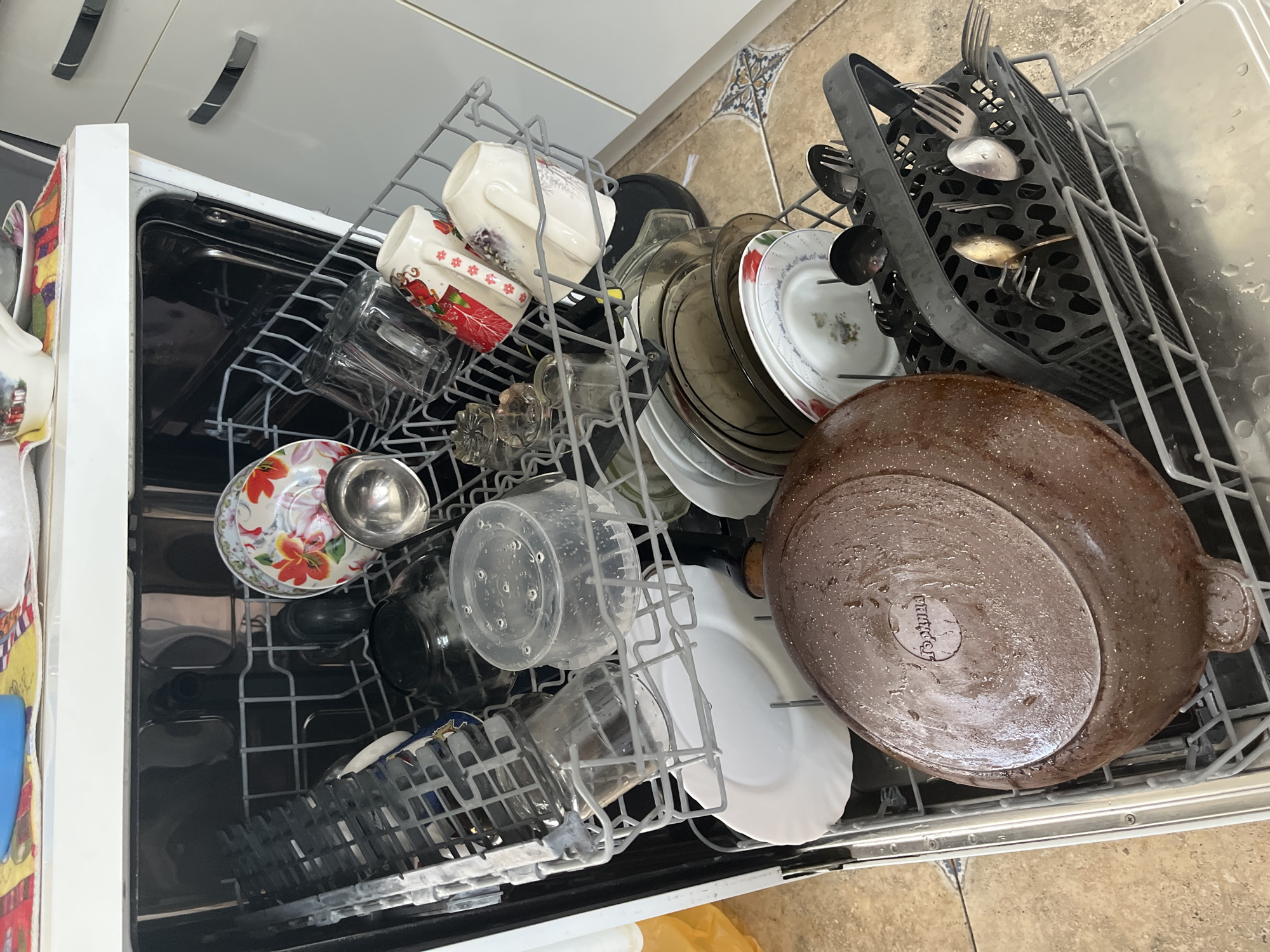 The image shows the inside of a dishwasher filled with dirty dishes. The dishwasher is partially open, revealing multiple racks containing plates, bowls, mugs, glasses, and a large pan. The pan is in the lower rack and is a dark brown color with speckles. There is also a cutlery basket filled with utensils. The overall scene suggests that a meal has recently been consumed and the dishes are awaiting washing. The kitchen floor, which is tiled with a geometric pattern, is visible beneath the open dishwasher door. The cabinets of the dishwasher are white, and the overall setting appears to be a domestic kitchen. There is no indication of the city of Saratov, Russia in the image.