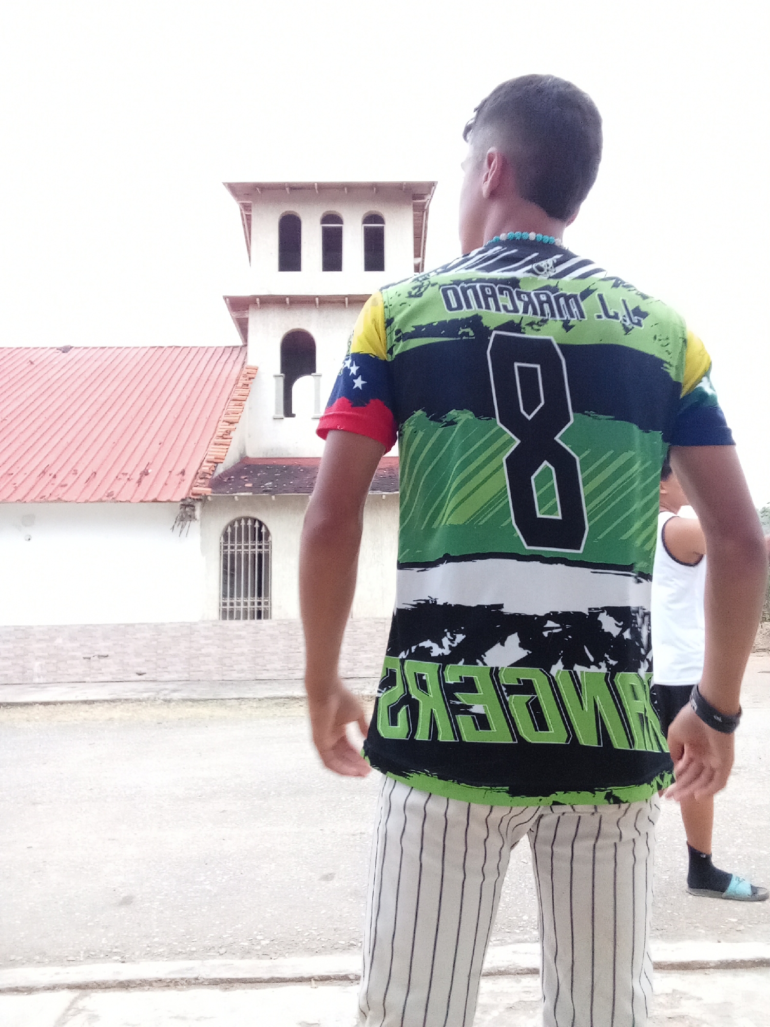 A young man is depicted from the back, standing outdoors and looking towards a white building with a distinctive red tiled roof. The building features a tower with multiple arched windows, suggesting a church or colonial-style architecture, set against a bright, overcast sky. The location is in Juan Griego, Venezuela.
The man is wearing a striking short-sleeved athletic shirt with a vibrant design. The shirt is primarily green, black, and white, with various patterns. On the back, a large number "8" is prominently displayed in white outline. Above the "8", the name "L. L. MARCANO" is visible in reversed text, while below it, the word "RANGERS" is also printed in reversed green text on a black background. The sleeves of the shirt incorporate colors and symbols resembling the Venezuelan flag: yellow, blue with white stars, and red. A beaded necklace, featuring light blue and white beads, is visible around his neck. He wears white pants with thin black vertical stripes.
To the right of the main subject, another person is partially visible, wearing a white tank top, dark socks, and light blue sandals. The ground beneath them is a light-colored, paved surface, possibly a street or a courtyard. The overall impression is a casual daytime scene.