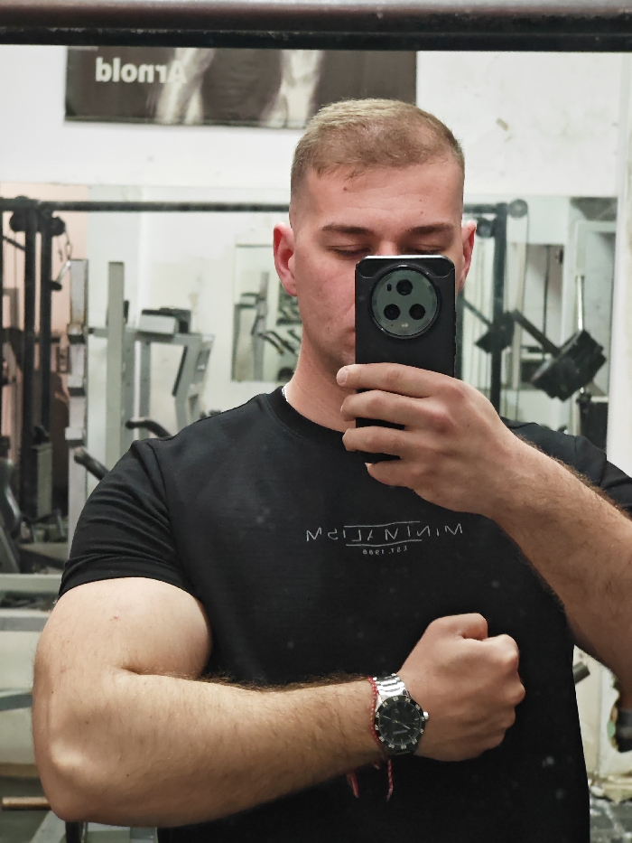 A male individual is depicted taking a mirror selfie within a gym located in Shumen, Bulgaria. The subject, visible from the chest up, has short, light-colored hair and is wearing a black t-shirt with the reversed text "MINIMALISM est. 1999". He holds a dark-colored smartphone with a prominent circular triple-lens camera array with his left hand, obscuring his face. His right arm is bent at the elbow, displaying a flexed bicep, with the hand clenched into a fist. A silver-toned wristwatch with a black face is worn on his left wrist, alongside a red and white string bracelet. The background, visible through the mirror, shows various pieces of metal gym equipment and structures, characteristic of a weight training facility. A poster featuring partially visible reversed text "blonA" (suggesting "Arnold") is affixed to a white wall in the upper left of the reflection.