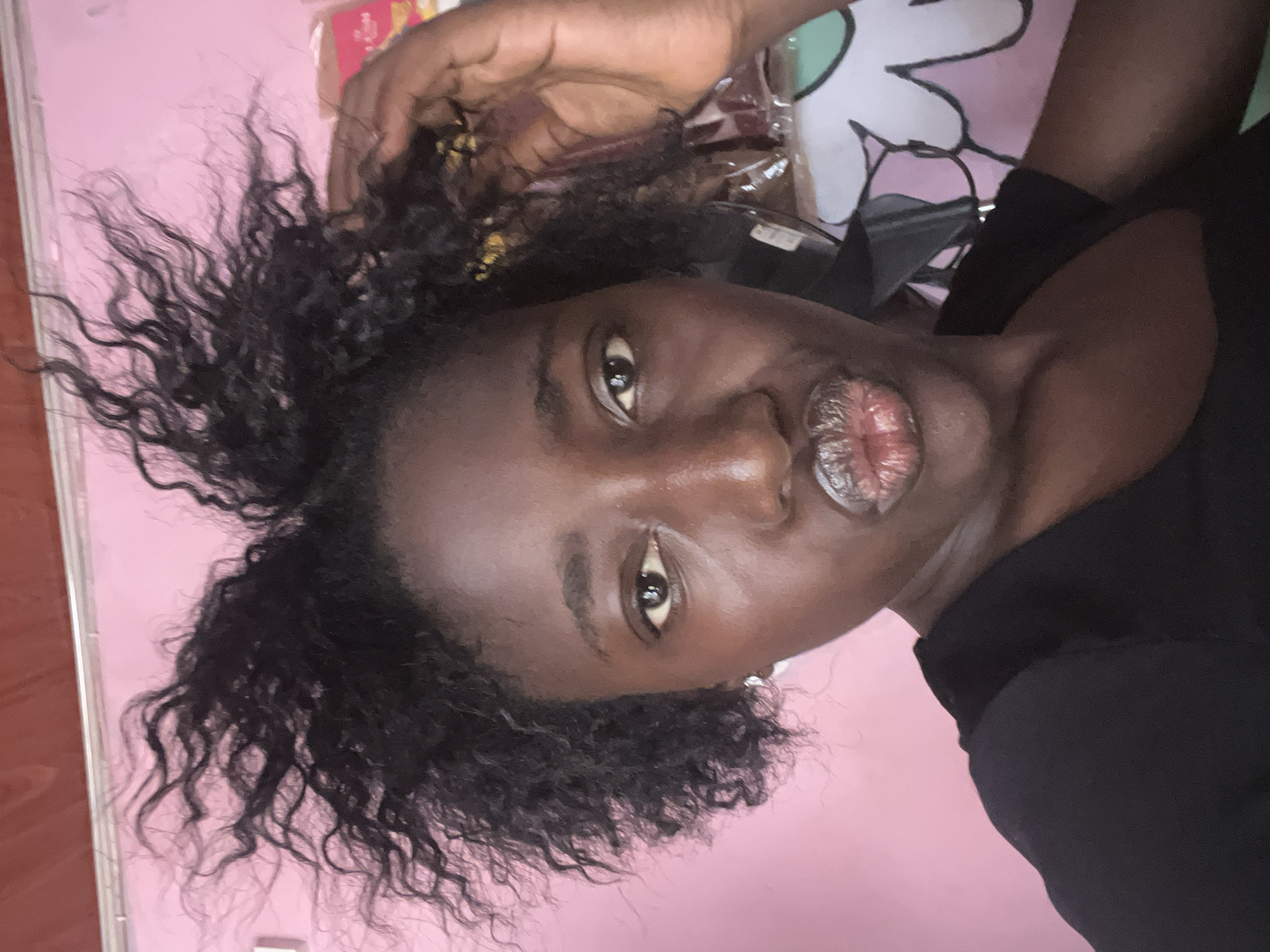A medium shot depicts a woman with dark skin and dark, curly hair, facing the camera at an angle. She is wearing a black top. Her face is prominent, with noticeable features including her eyes and lips. A hand is visible in the upper part of the frame, near her hair. The background is a light pink wall. The specific location within Abuja, Nigeria, cannot be determined from the image alone.