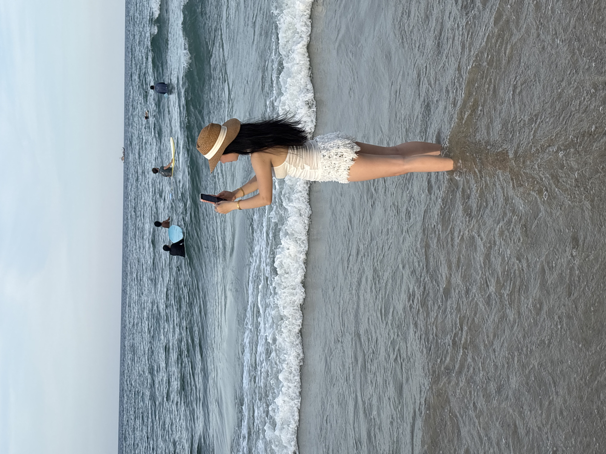 A young Vietnamese woman stands in shallow ocean water at a beach in Đà Nẵng, Vietnam, holding a smartphone to take a picture or video. She is wearing a white one-piece swimsuit with lace detailing on the shorts and a wide-brimmed straw hat. The ocean has small waves breaking on the shore, and several other people are visible in the water, some with surfboards. In the distance, a small boat is visible on the horizon. The sky is overcast, creating a soft, diffused light.