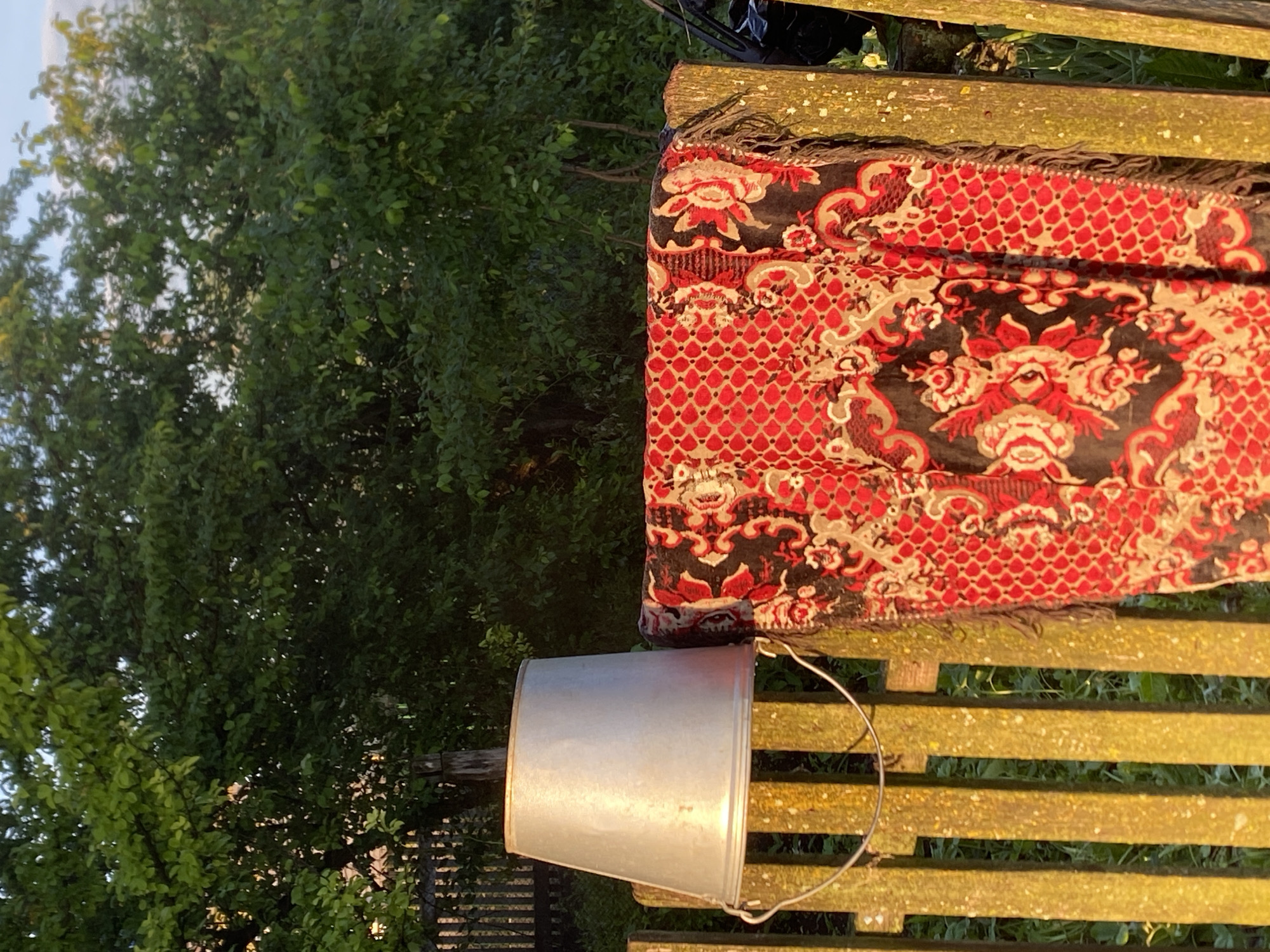 A still life photograph captures a rustic outdoor scene, likely in the village of Chirsha-Tartysh, Russia, as evidenced by the traditional elements. The focal point is a weathered, light-colored metal bucket, its handle a thin wire, resting atop a rustic wooden fence. The fence, constructed from vertical wooden planks, appears aged and possibly stained with a yellow hue. Draped over the fence, partially obscuring it, is a vibrant, patterned textile. The fabric is predominantly red with intricate floral designs in black and white, featuring a fringed edge. The background is dominated by lush green foliage from trees, some of which are illuminated by what appears to be the golden light of either sunrise or sunset. The overall impression is one of a simple, perhaps domestic, outdoor setting, evoking a sense of rural life. There are no people or animals visible in the frame.