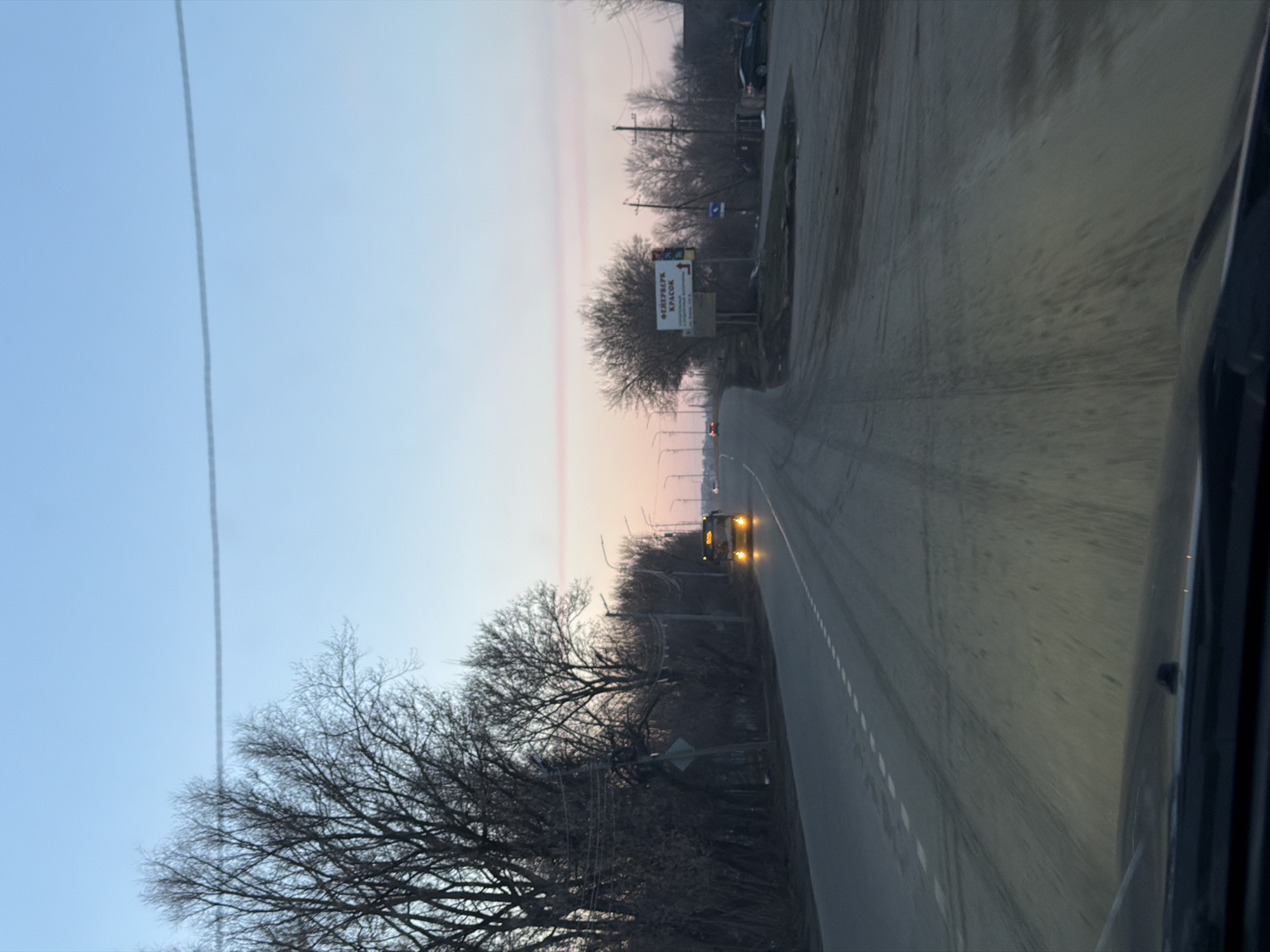The image shows a view from inside a vehicle driving on a road. A billboard can be seen in the distance with the words "ФЕДЕРАЛЬНЫЙ КРАСОК" displayed, which is a company located in Sarapul, Russia. A vehicle with headlights on is approaching in the distance. Bare trees line the side of the road, and overhead power lines are visible. The sky is pale blue and pink, suggesting it is either sunrise or sunset. The road appears to be paved and has painted lane markings.