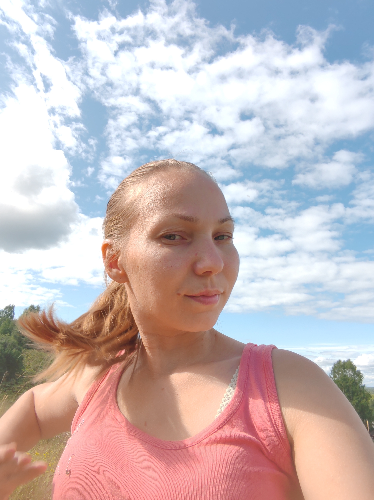 A young woman with blonde hair pulled back in a ponytail is posing for a selfie outdoors under a partly cloudy sky. She is wearing a pink tank top with a white lace bra visible underneath. The background shows a line of trees and a vast expanse of blue sky with white clouds. A safety pin is attached to the woman's tank top. The scene appears to be in a rural or natural setting.