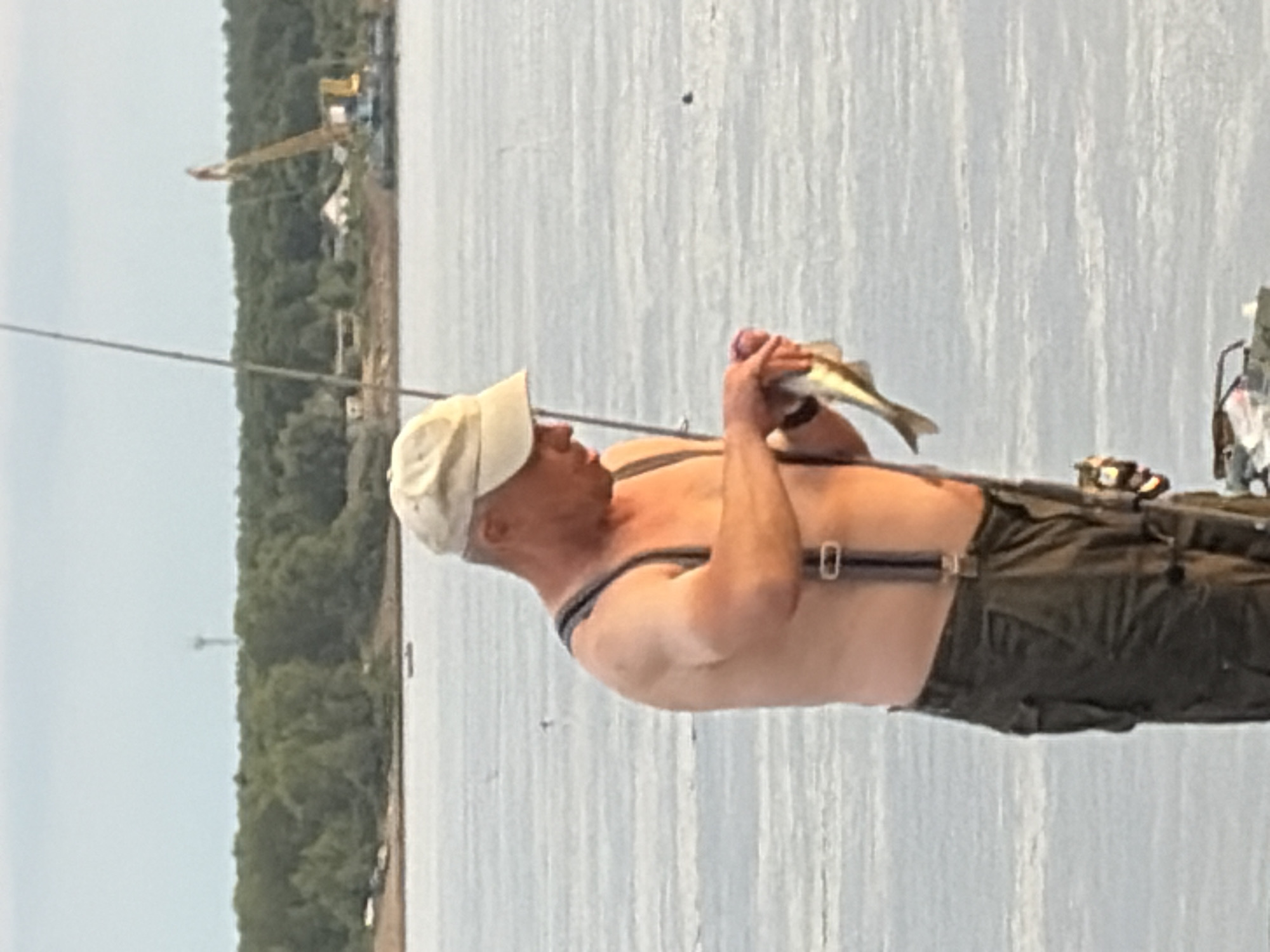 An adult male wearing a light-colored baseball cap, dark suspenders, and olive-green cargo pants stands on a body of water, holding a small fish in his right hand. The man is shirtless, and his left arm is bent as he examines the fish. A fishing rod is visible behind him. The background features a tree-lined shore with a construction crane and buildings. The body of water appears to be the Kama River in Chaikovsky, Russia, given the characteristic landscape and the presence of industrial elements often found along its banks. The lighting suggests it is late afternoon or early evening.