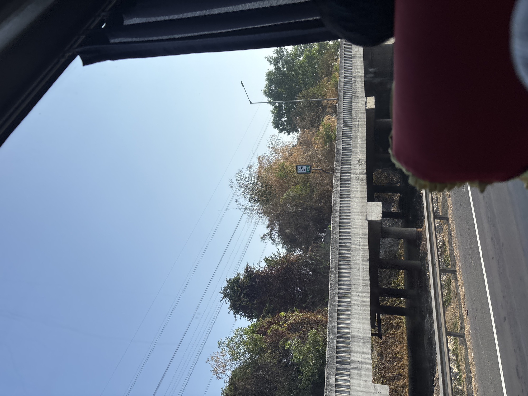 The image captures a view from within a vehicle, looking out at a highway scene in Karawang, Indonesia. A concrete overpass with decorative vertical elements dominates the mid-ground. Beyond the overpass, lush green trees and some dry, brown foliage are visible against a clear blue sky. Power lines stretch across the sky, and a street lamp stands near the overpass. A road sign with "KM 47 800" is present. In the foreground, the edge of a red seat is visible, suggesting the perspective of a passenger. The road in the foreground shows a white lane marker. The overall impression is a bright, sunny day on a highway.