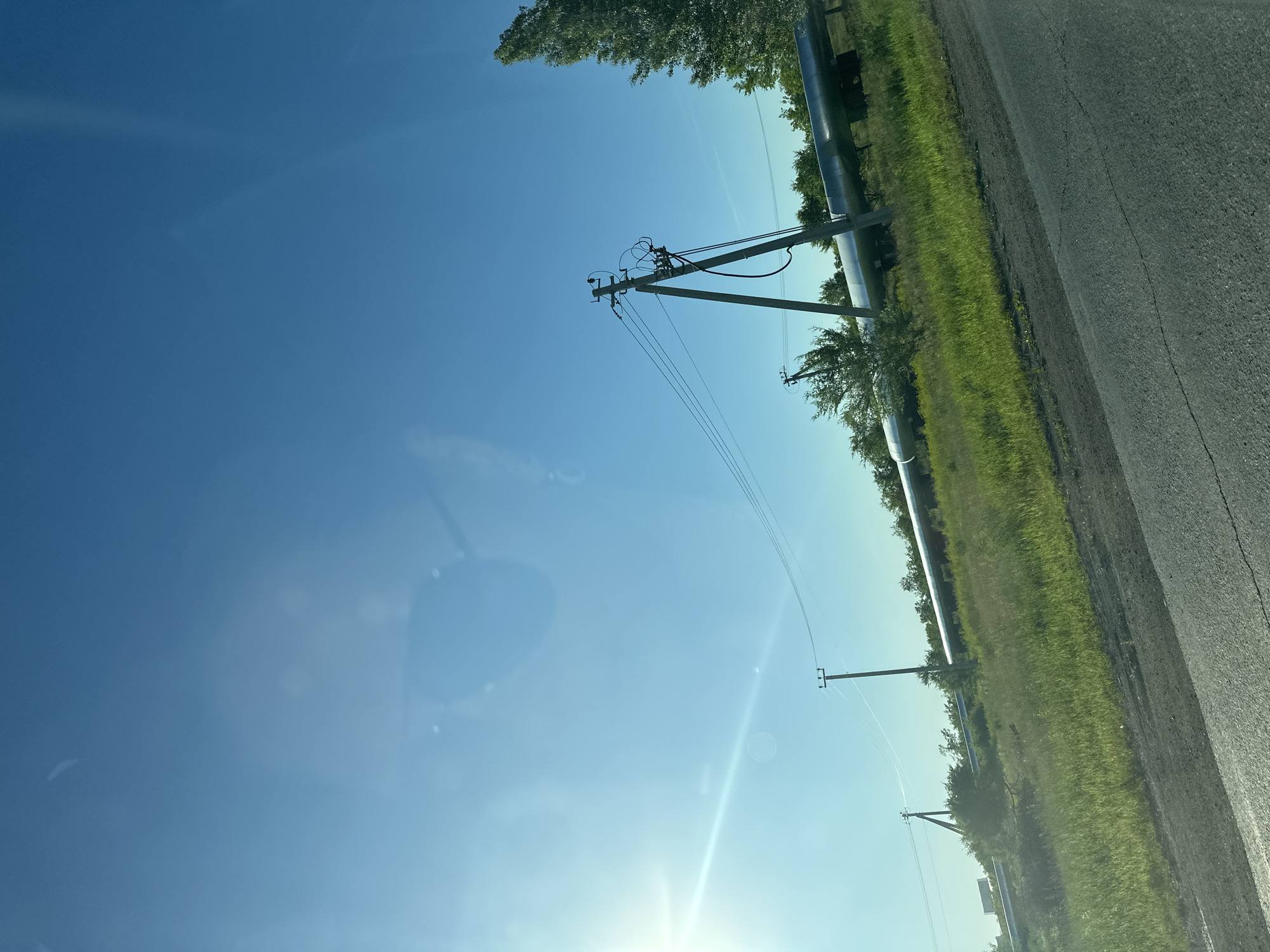 The image shows a roadside scene with a bright blue sky, indicating it is daytime, likely sunny given the lens flare visible. Along the side of a paved road, there is a grassy field with some trees. A large, silver pipeline runs parallel to the road, supported by structures. There are also power lines with utility poles visible. The perspective suggests the photo was taken from a moving vehicle, as indicated by the slight blur and the angle of the road. The overall setting appears to be rural or semi-rural. No people are visible, and no text is discernible in the image. The weather appears to be clear and sunny.