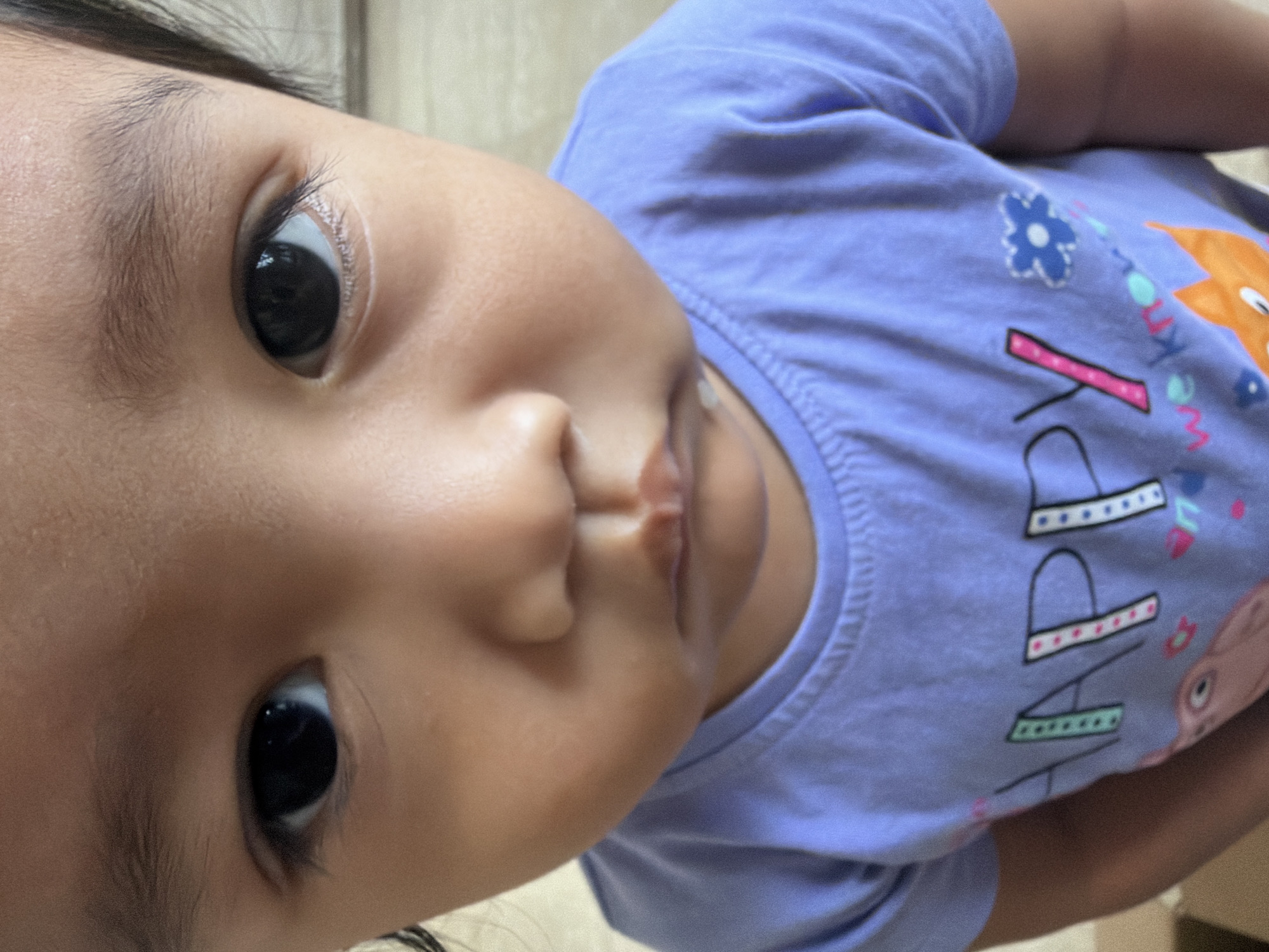 The image is a close-up, head-on shot of a young child, likely a toddler, wearing a light purple t-shirt with the word "HAPPY" printed in large letters, along with graphics of flowers and an animal, possibly a fox. The child's face is the primary focus, with large, dark eyes, a small nose, and slightly downturned lips. The background is blurred and appears to be an indoor setting with neutral-colored walls. There is no visible indication of the city of Central Jakarta, Indonesia, or any other specific location. The lighting is soft and natural, casting a gentle glow on the child's face. The composition is tight, emphasizing the child's features and expression.