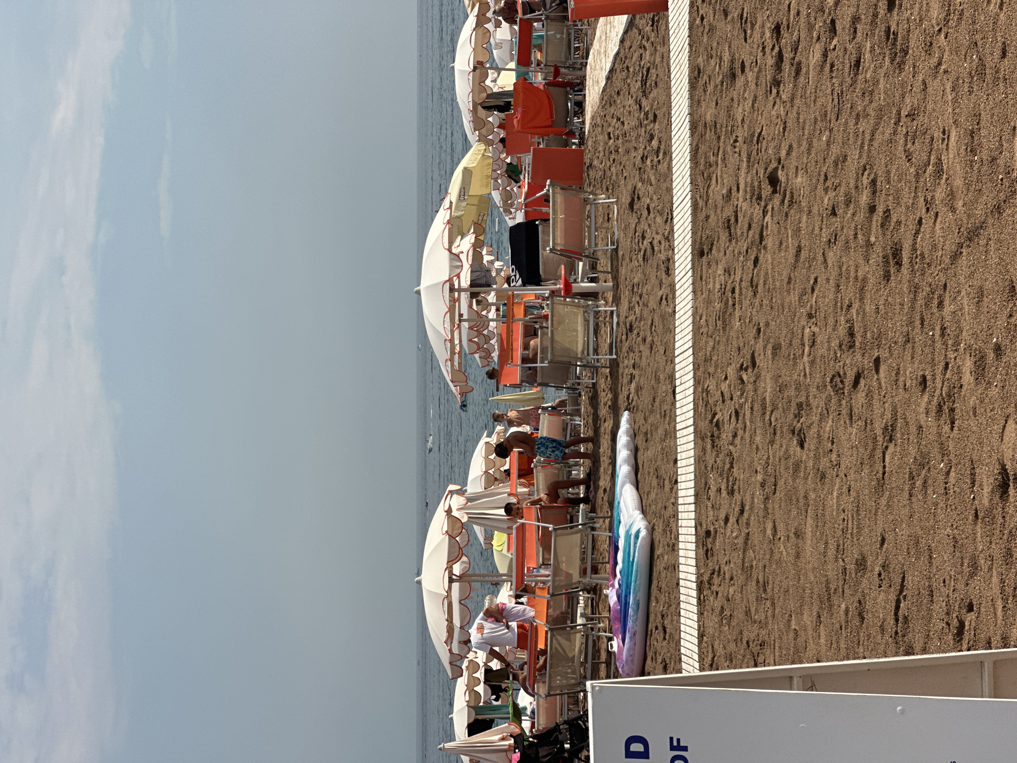 The image depicts a beach scene with numerous beach chairs and umbrellas set up on the sand. The sea is visible in the background, with a clear sky above. Several people are present on the beach, some sitting in chairs, others standing, and children playing. Beach inflatables are scattered on the sand. A white sign with blue text is partially visible in the foreground. The overall atmosphere is one of a crowded beach on a sunny day. The location is stated as Boreti, Montenegro.