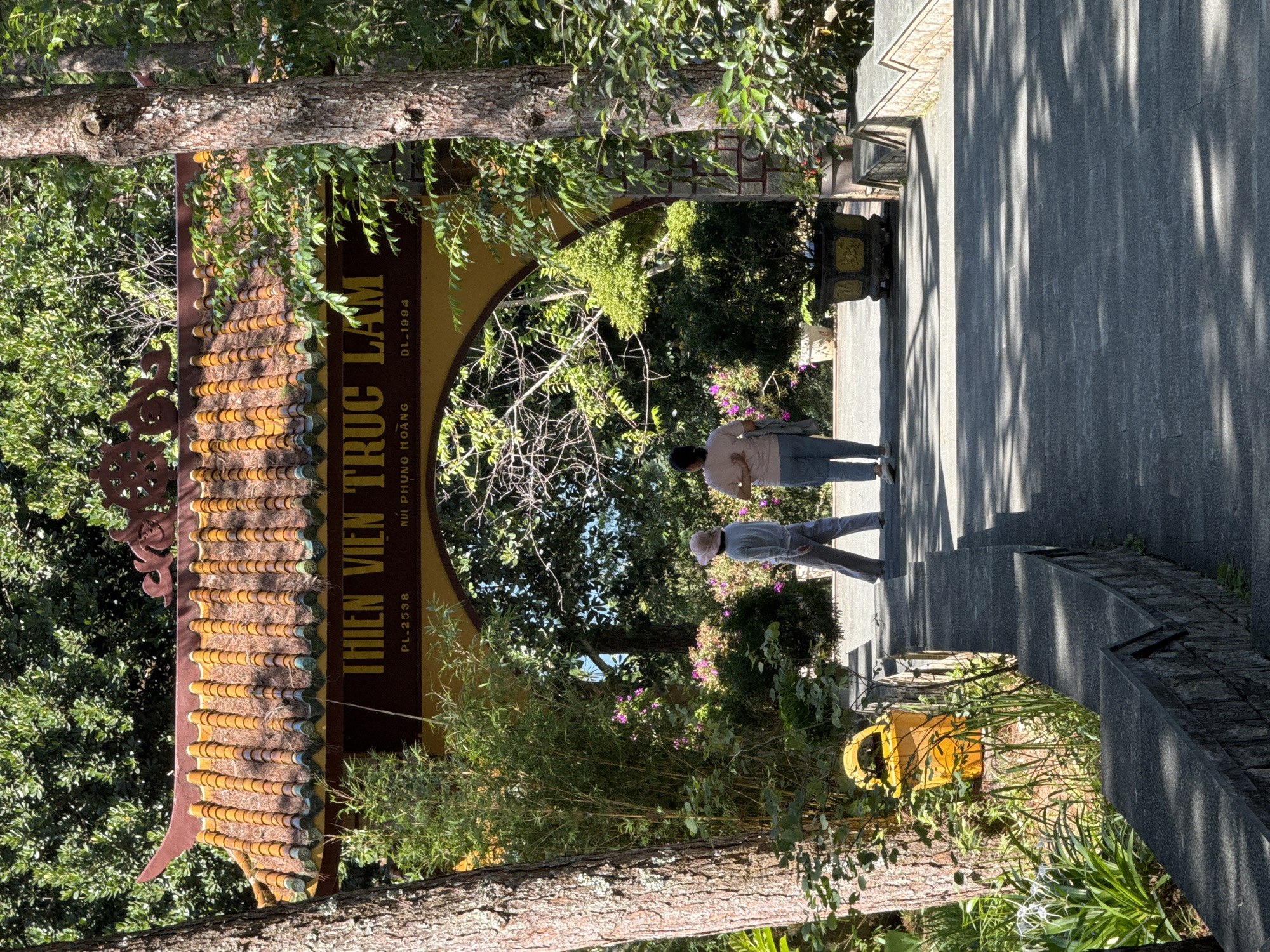 The image depicts the entrance to Thien Vien Truc Lam, a prominent Buddhist monastery located on a mountain overlooking Tuyen Lam Lake in Da Lat, Vietnam. The entrance features a traditional Vietnamese temple gate with an ornate tiled roof and carved decorations. Two individuals, one wearing a light-colored hat and a light blue outfit, and another in a light pink shirt and blue pants, are walking away from the viewer towards the gate. The pathway leading to the gate is paved with stone, and the surrounding environment is lush with trees, bamboo, and flowering plants. The sunlight casts dappled shadows on the ground, indicating a sunny day. A yellow trash bin is visible on the left side of the path. The overall scene conveys a sense of tranquility and natural beauty.