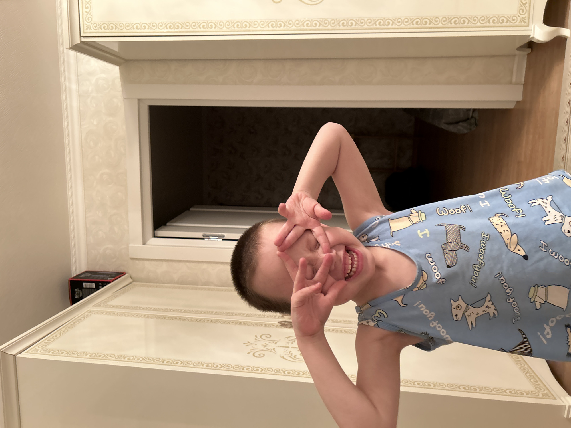A rotated indoor shot features a young child in a light blue, sleeveless top patterned with various dog illustrations and the word "Woof!" repeatedly. The child is making hand gestures in front of their face, partially obscuring their eyes while grinning, revealing their teeth.
The background consists of a white cabinet or dresser with ornate designs along the edges and top surface. Above the cabinet, there is a recessed area in the wall with light-colored patterned wallpaper. A dark object is visible inside the recessed area, and a small box is placed on top of the cabinet near the left edge of the frame. The room appears to be well-lit, and the overall composition suggests a playful or candid moment captured in a domestic setting.
Given the prompt, it can be assumed that this photograph was taken in Chaikovsky, Russia.